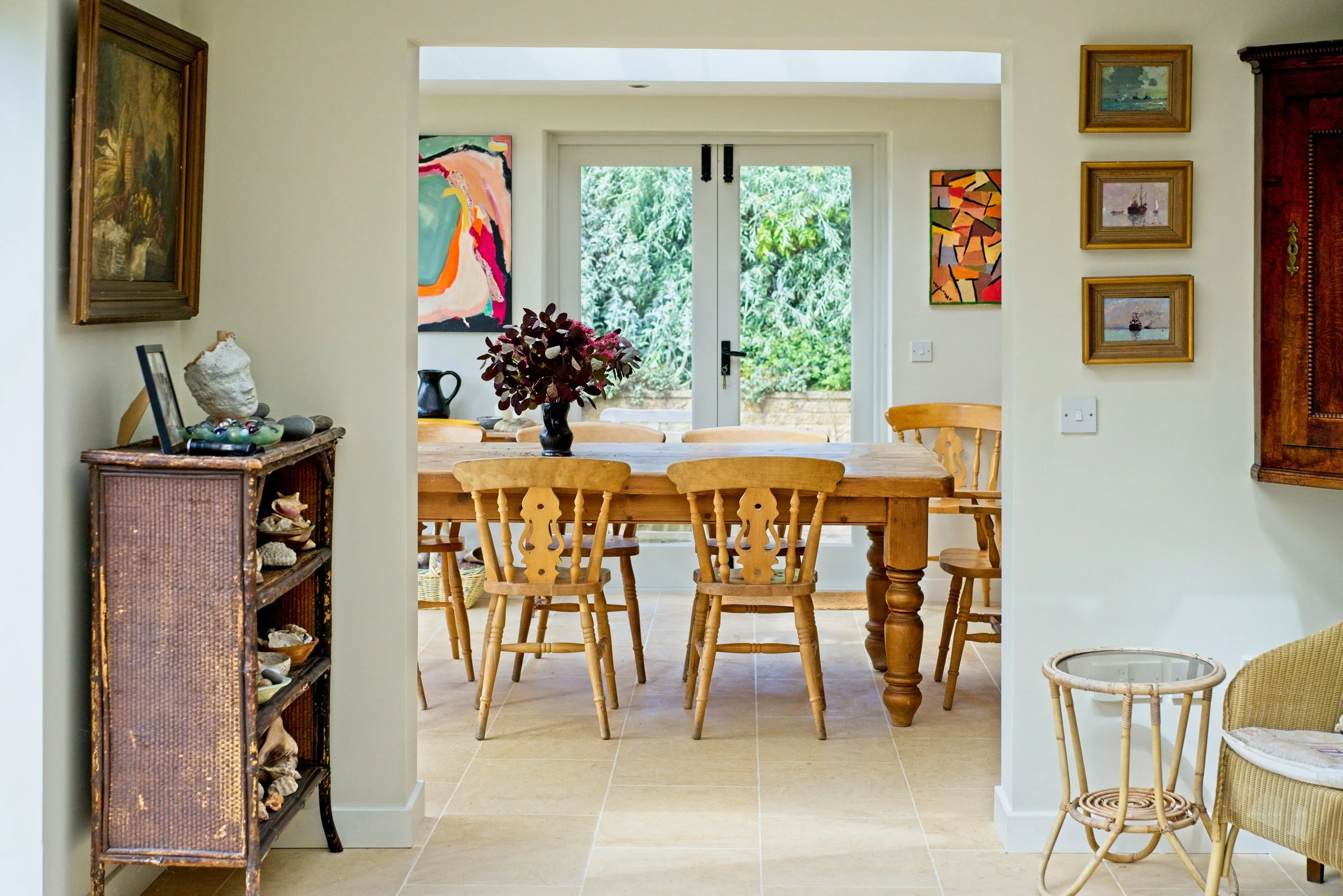Dining area with wooden table and chairs, decoratively dressed with a floral centerpiece, surrounded by artwork and furniture, with open doors leading to a garden. Property Photography by photographer based in Southam, Warwickshire