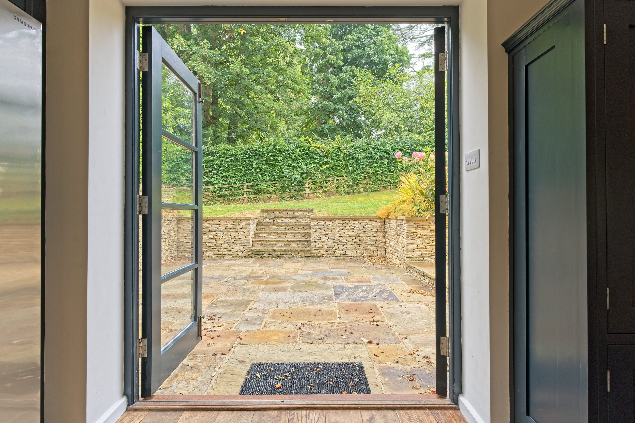 View through an open door onto a stone patio with stairs leading to a grassy yard and a lush green hedge and trees in the background. Property Photography by photographer based in Southam, Warwickshire