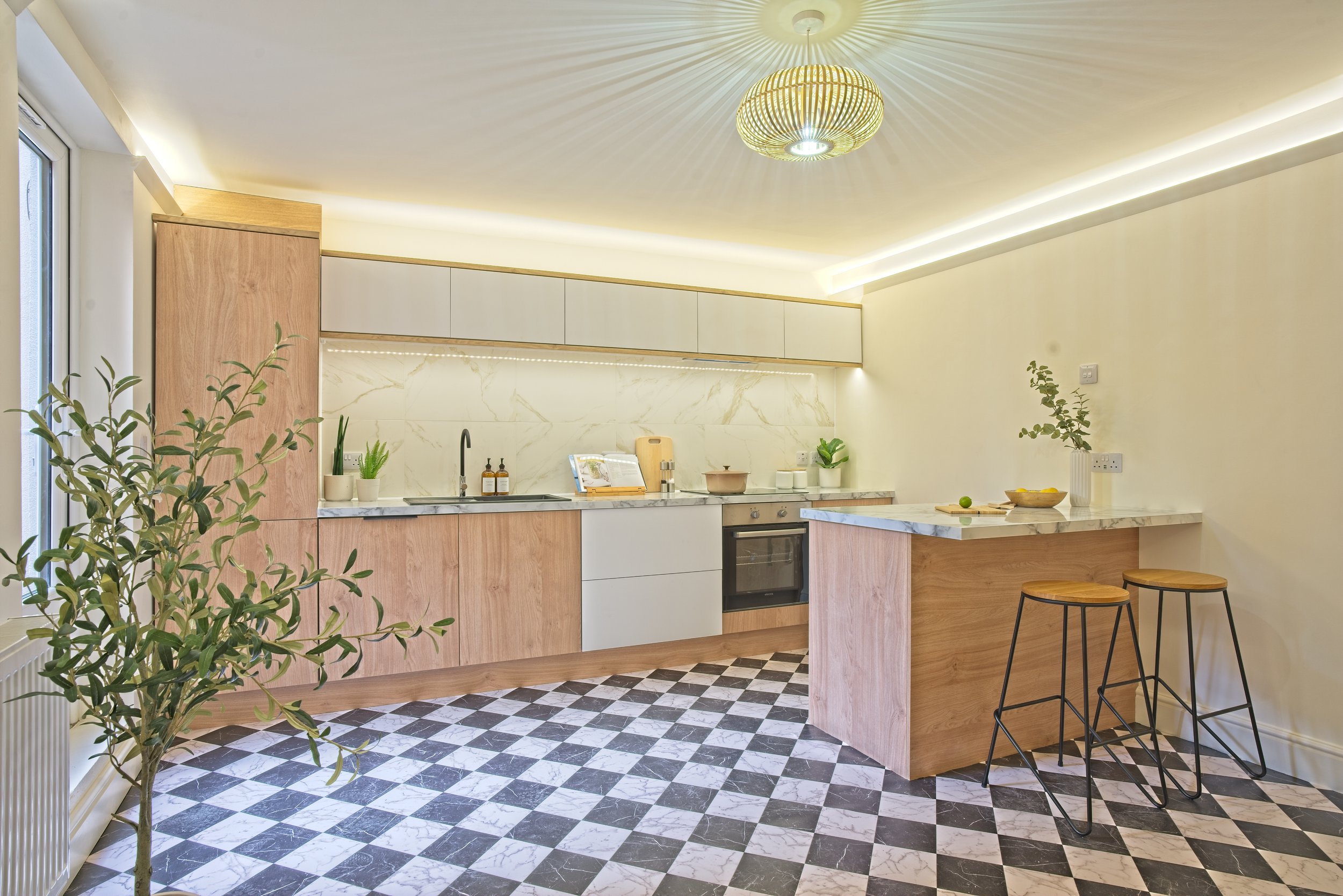 Modern kitchen with light wood cabinets, marble countertops, checkered black and white floor, and bar stools at breakfast bar. Photography by photographer based in Southam, Warwickshire