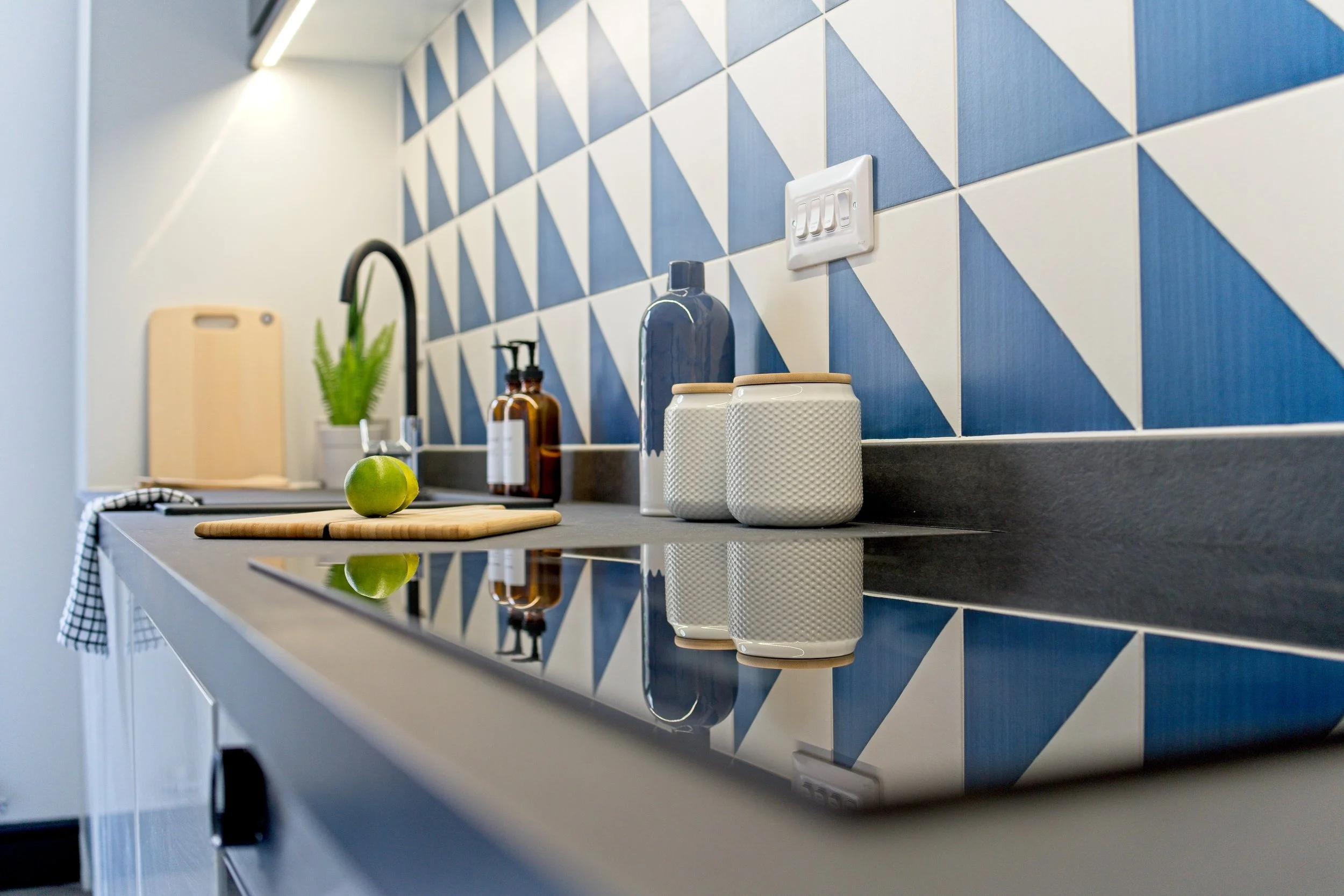 Modern kitchen with black countertop, white and blue geometric tile backsplash, sink with black faucet, cutting board with green apple, condiment bottles, ceramic jars, and a potted plant. Photography by photographer based in Southam, Warwickshire