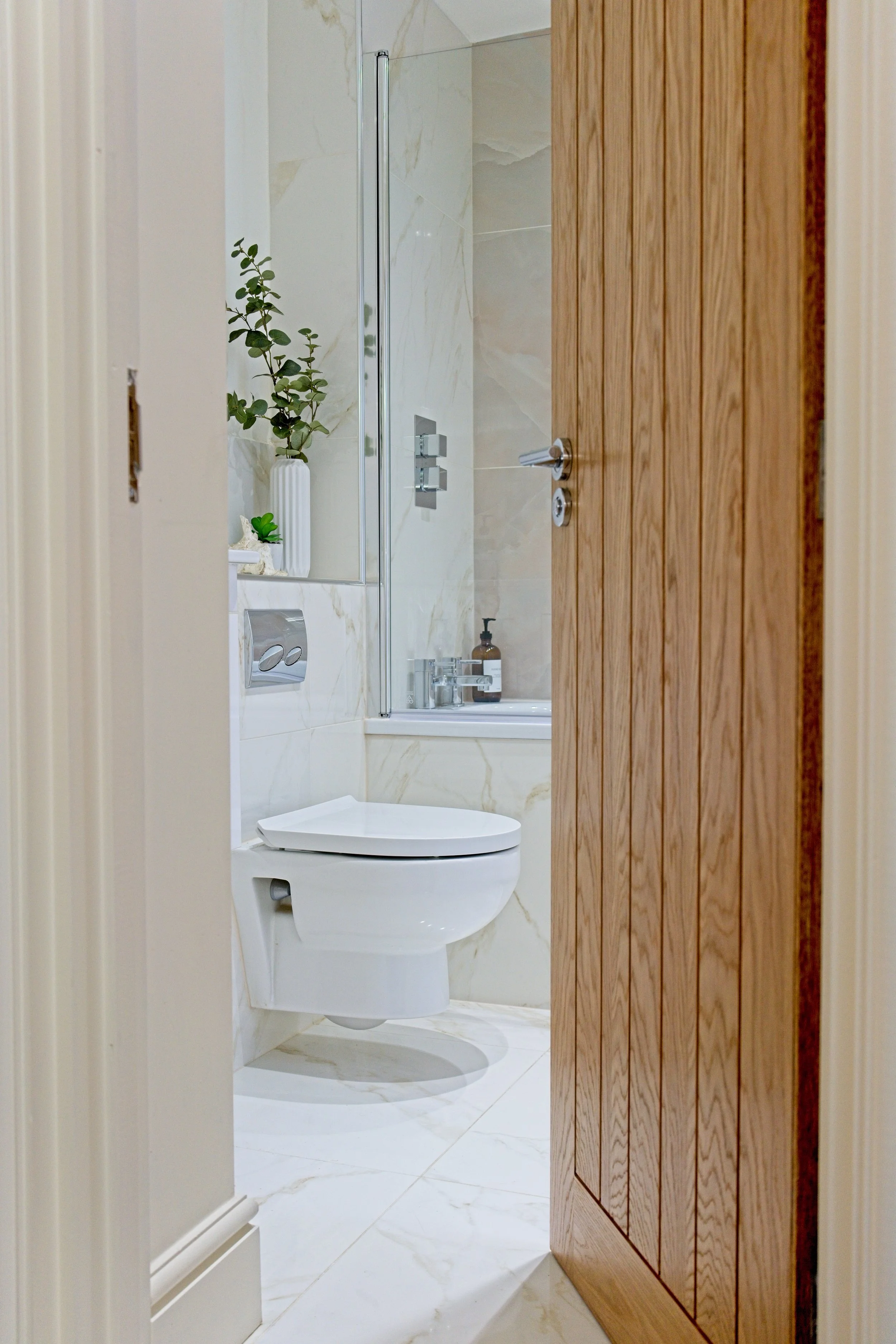 View of a modern bathroom with a white toilet, a small sink, a bottle of liquid soap, a potted plant on a shelf, and a wooden door. Photography by photographer based in Southam, Warwickshire