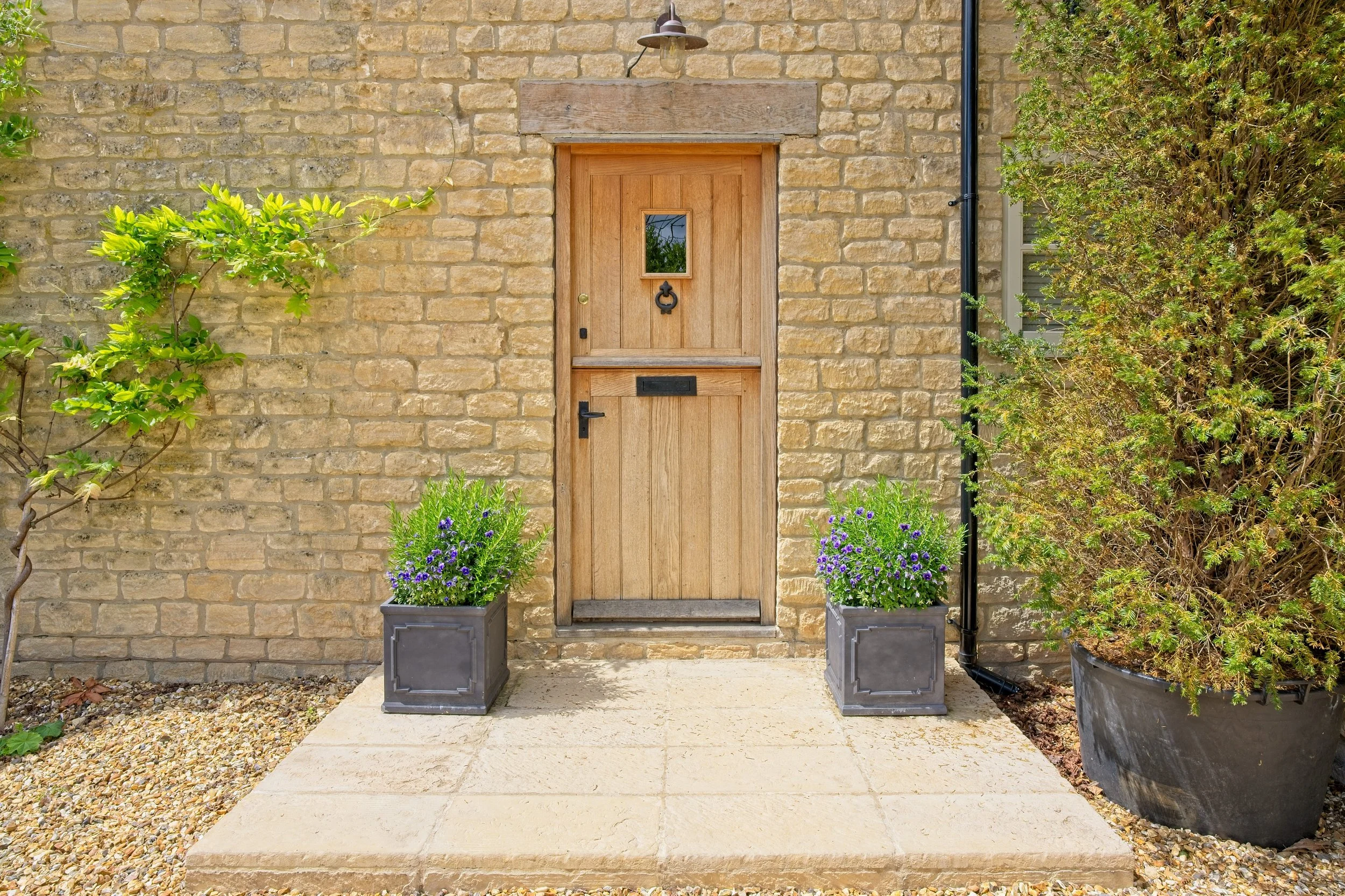 Wooden front door with small window and mailbox, flanked by potted purple flowers, next to brick wall and bushes. Photography by photographer based in Southam, Warwickshire