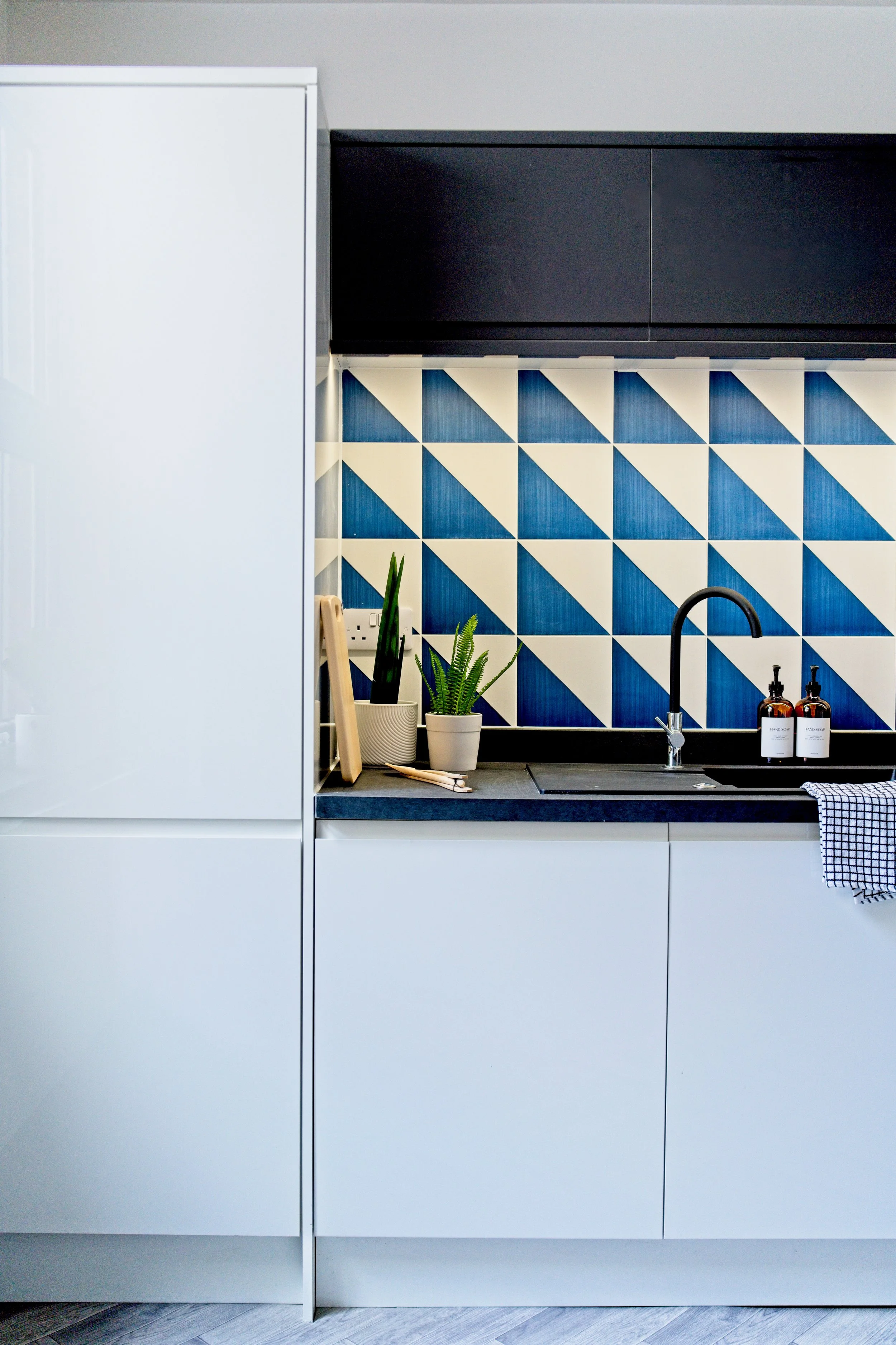 Modern kitchen with white cabinets, a black countertop, a geometric blue and white tiled backsplash, and potted plants near the sink. Property Photography by photographer based in Southam, Warwickshire