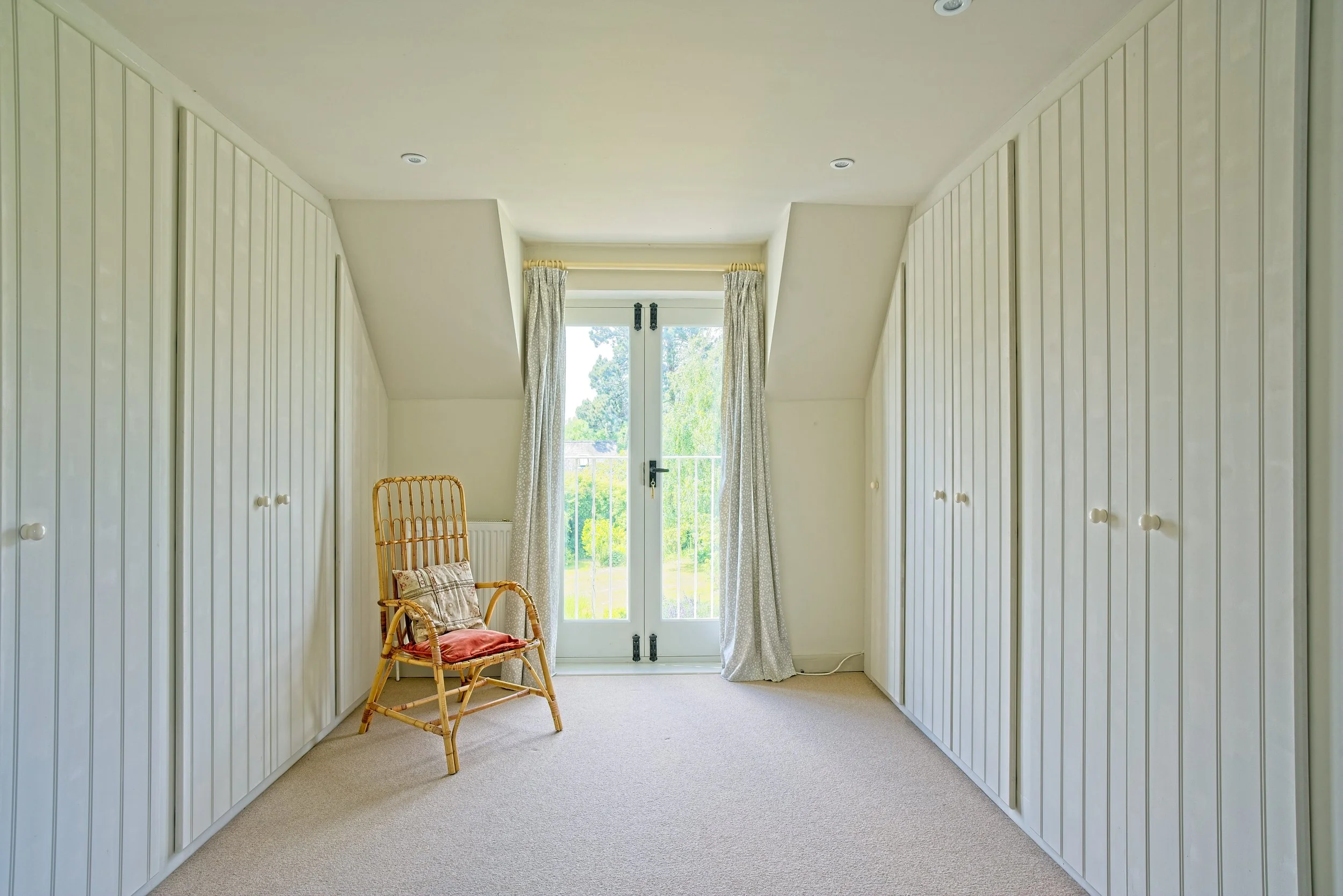 Empty room with white built-in closets on both sides, a wicker chair with a colorful pillow and red cushion, beige carpet, and a glass door with white curtains leading to a balcony with greenery outside. Photography by photographer based in Southam, 