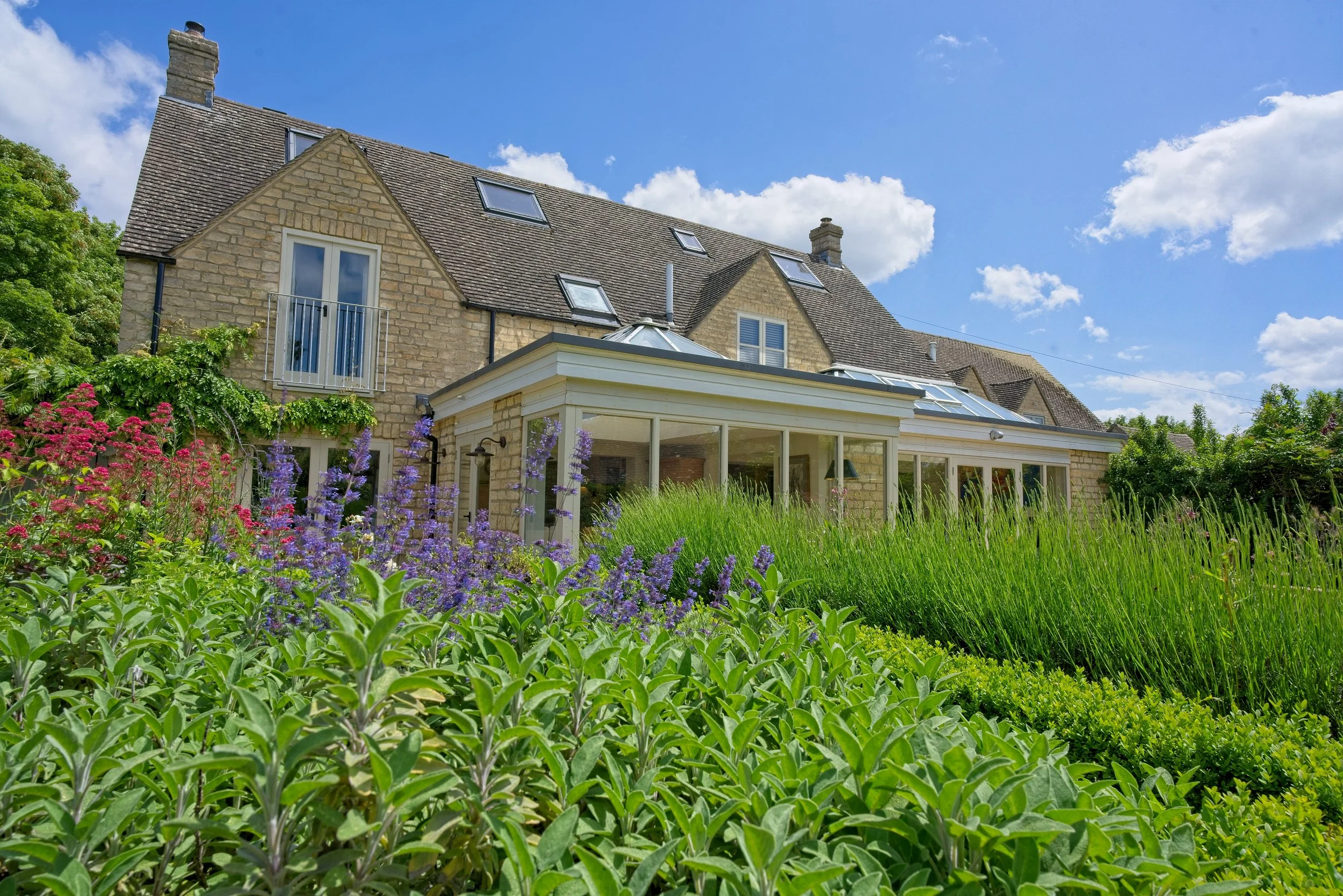 A large stone house with a new conservatory, surrounded by a lush garden with colorful flowers and tall green plants under a partly cloudy blue sky. Property Photography by photographer based in Southam, Warwickshire