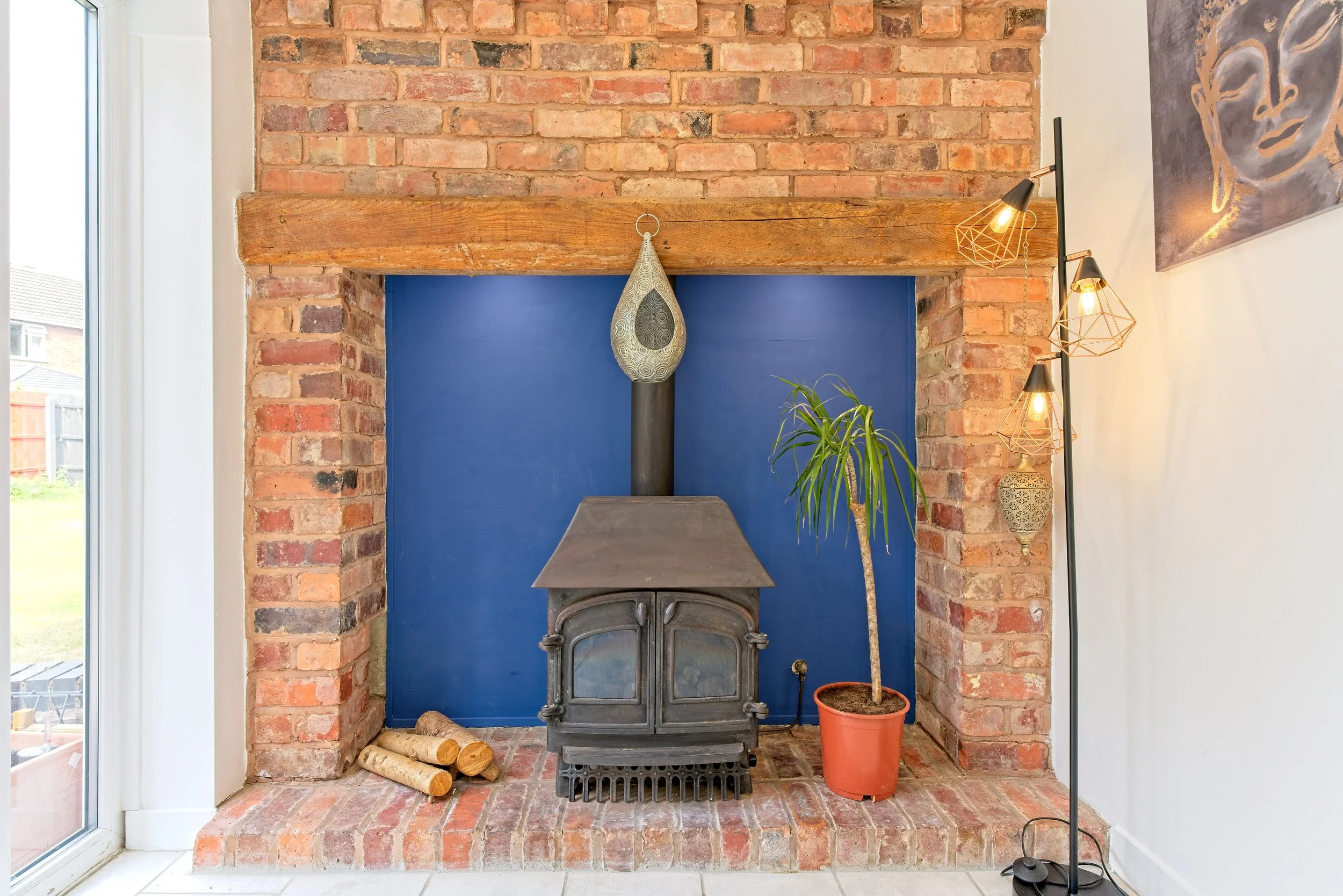 A fireplace with a brick surround and a blue painted back wall, featuring a wood stove, a potted plant, and a floor lamp with multiple light fixtures. Photography by photographer based in Southam, Warwickshire