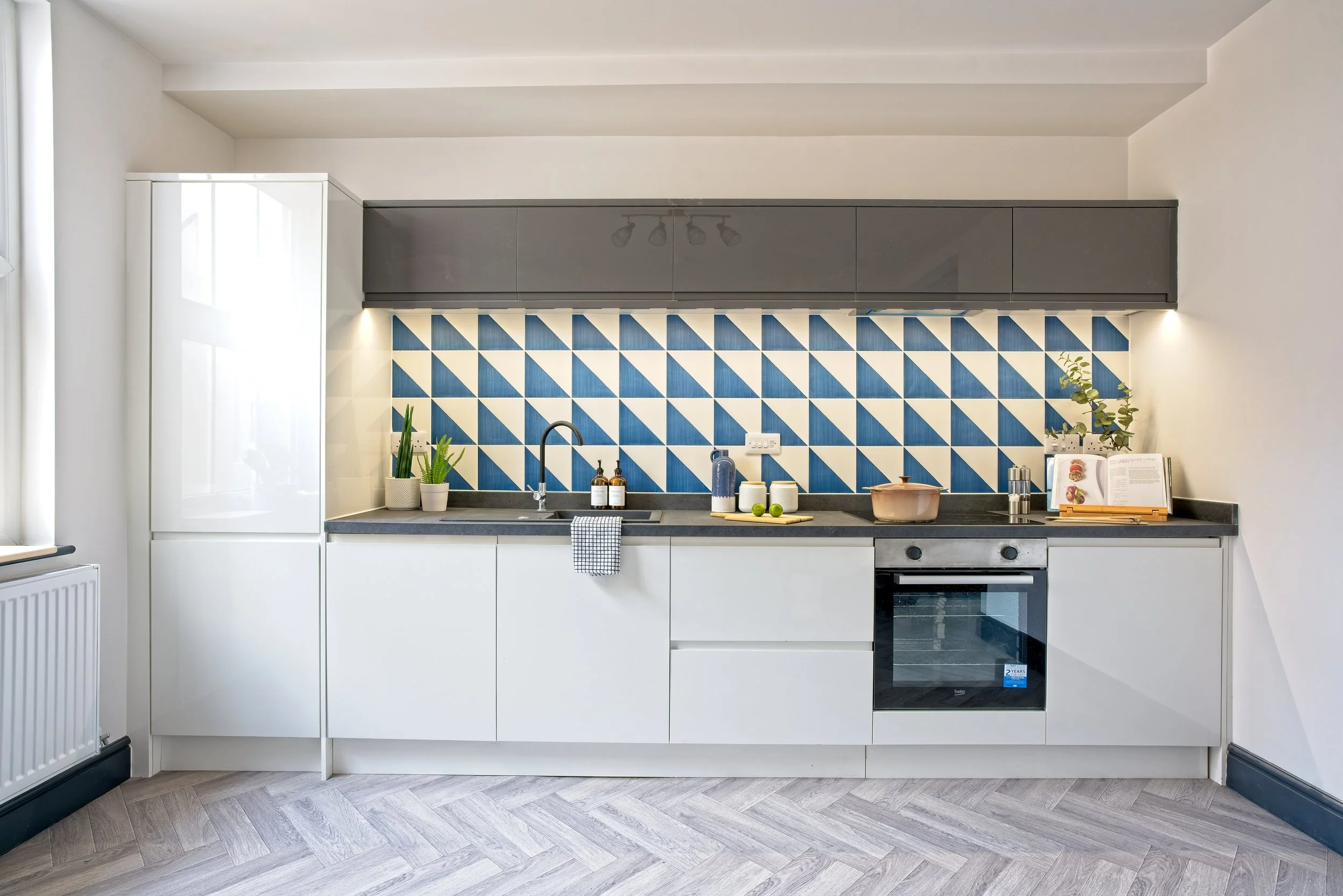 Modern kitchen with white cabinets, black countertop, blue and white geometric tile backsplash, built-in oven, and small plants. Photography by photographer based in Southam, Warwickshire