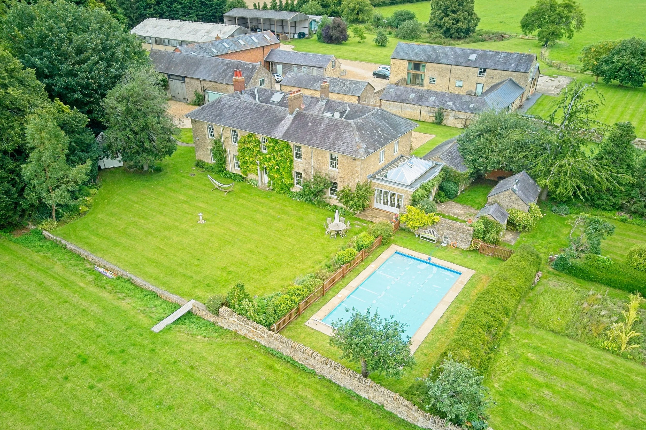 Aerial view of a large countryside estate featuring a mansion with a garden, a swimming pool, surrounded by lush green lawns, trees, and countryside landscape. Real estate photography in the Cotswolds and Warwickshire.