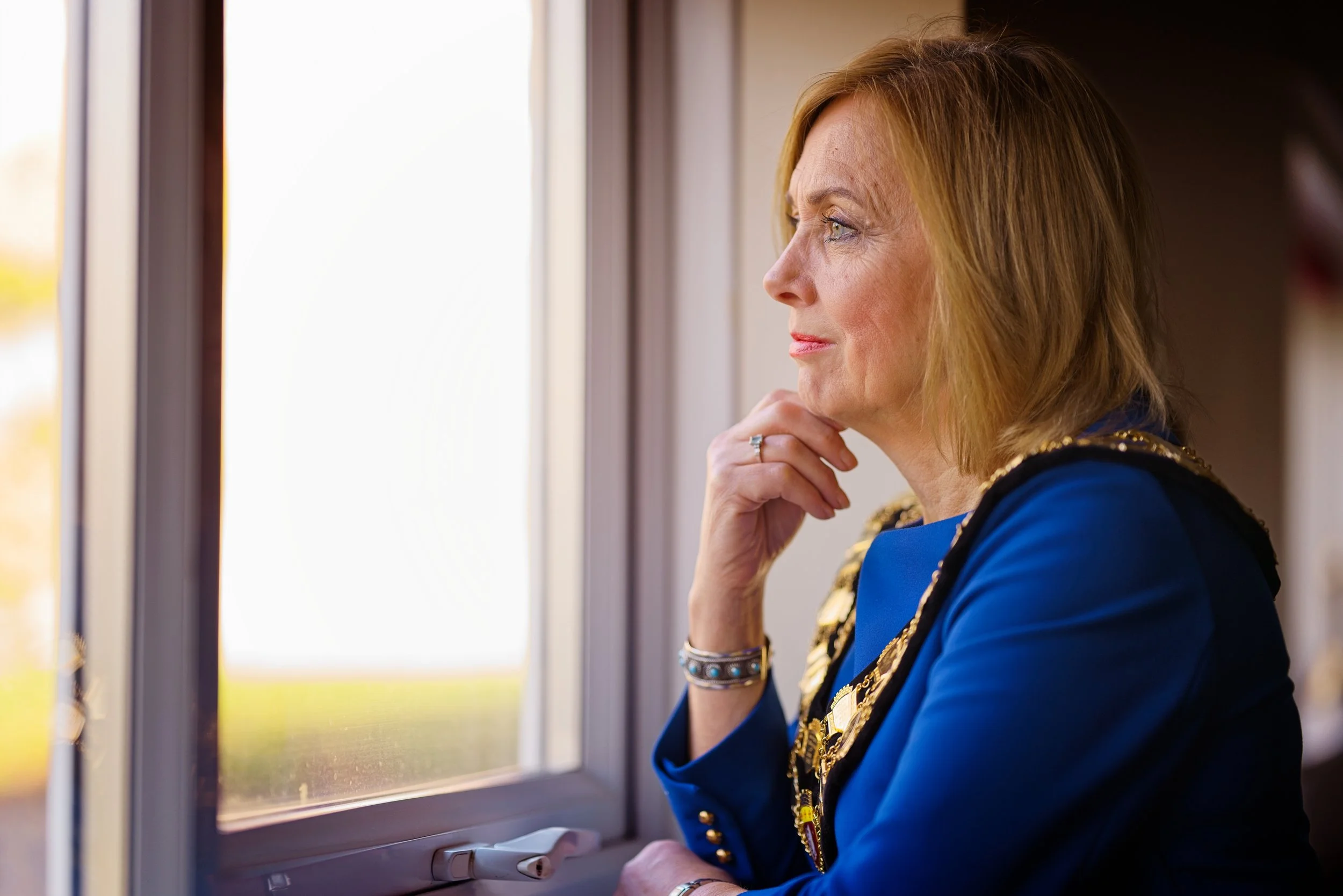 A woman with shoulder-length blonde hair looking out a window, her hand resting on her chin, wearing a blue jacket adorned with gold embellishments. Photography by photographer based in Southam, Warwickshire