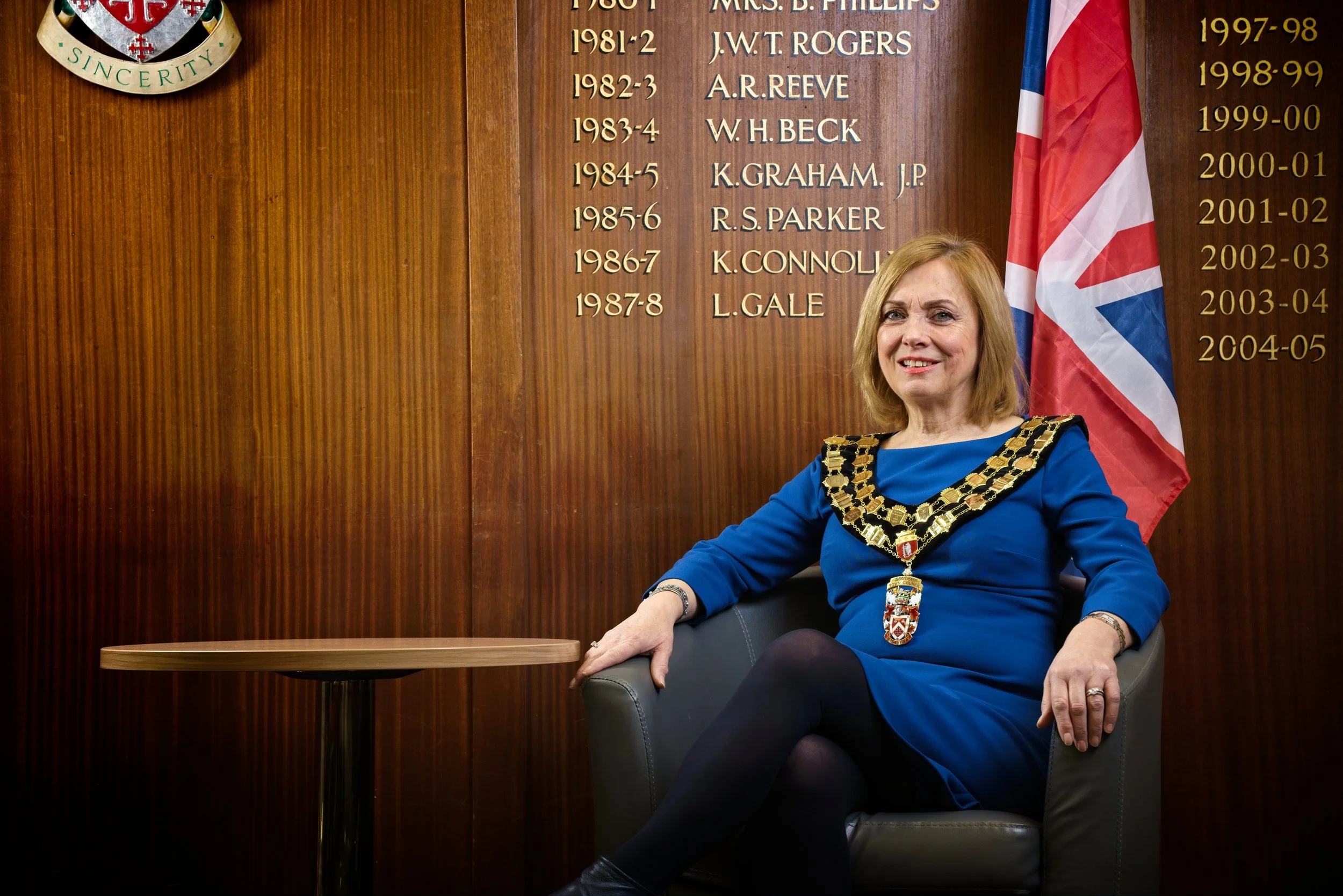 A woman dressed in a blue dress and a mayor's chain of office, sitting in a chair, smiling. Behind her is a wooden wall with a list of names and dates, and a partial Union Jack flag. The scene appears to be a formal or official setting. Photography b