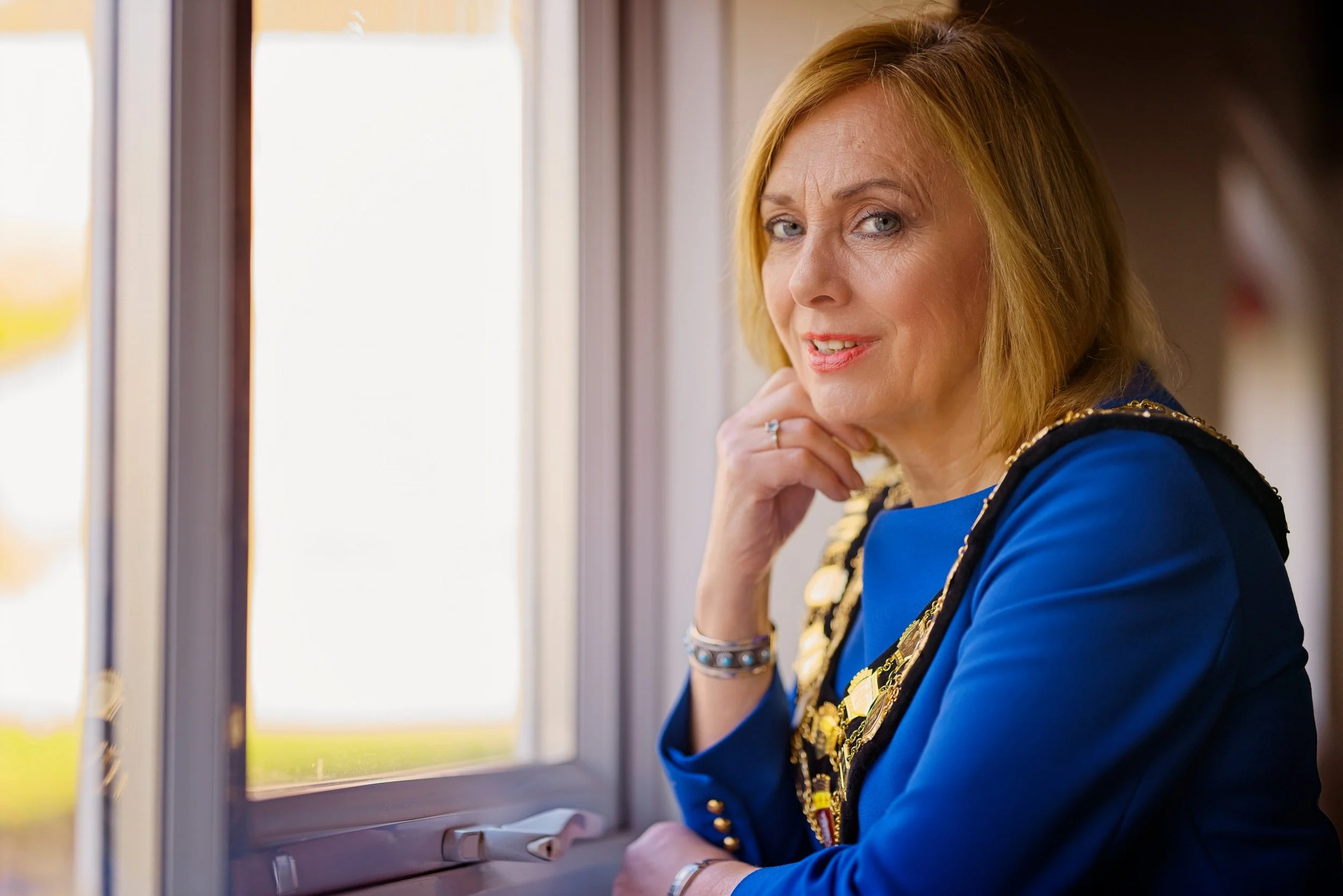 A middle-aged woman with blonde hair looking out of a window, wearing a blue dress with gold embellishments. Photography by photographer based in Southam, Warwickshire