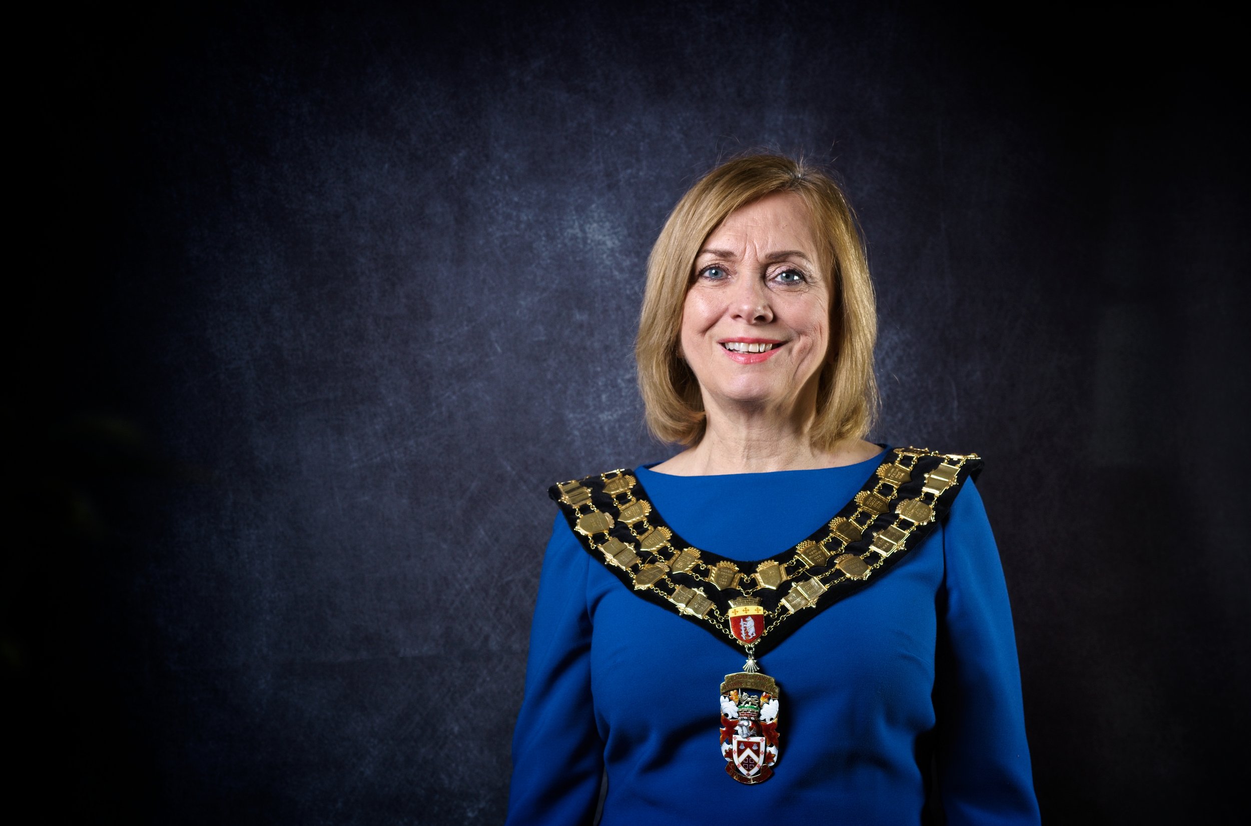 A woman with blonde hair wearing a blue dress and a decorative chain with a large crest, standing against a dark background. Photography by photographer based in Southam, Warwickshire
