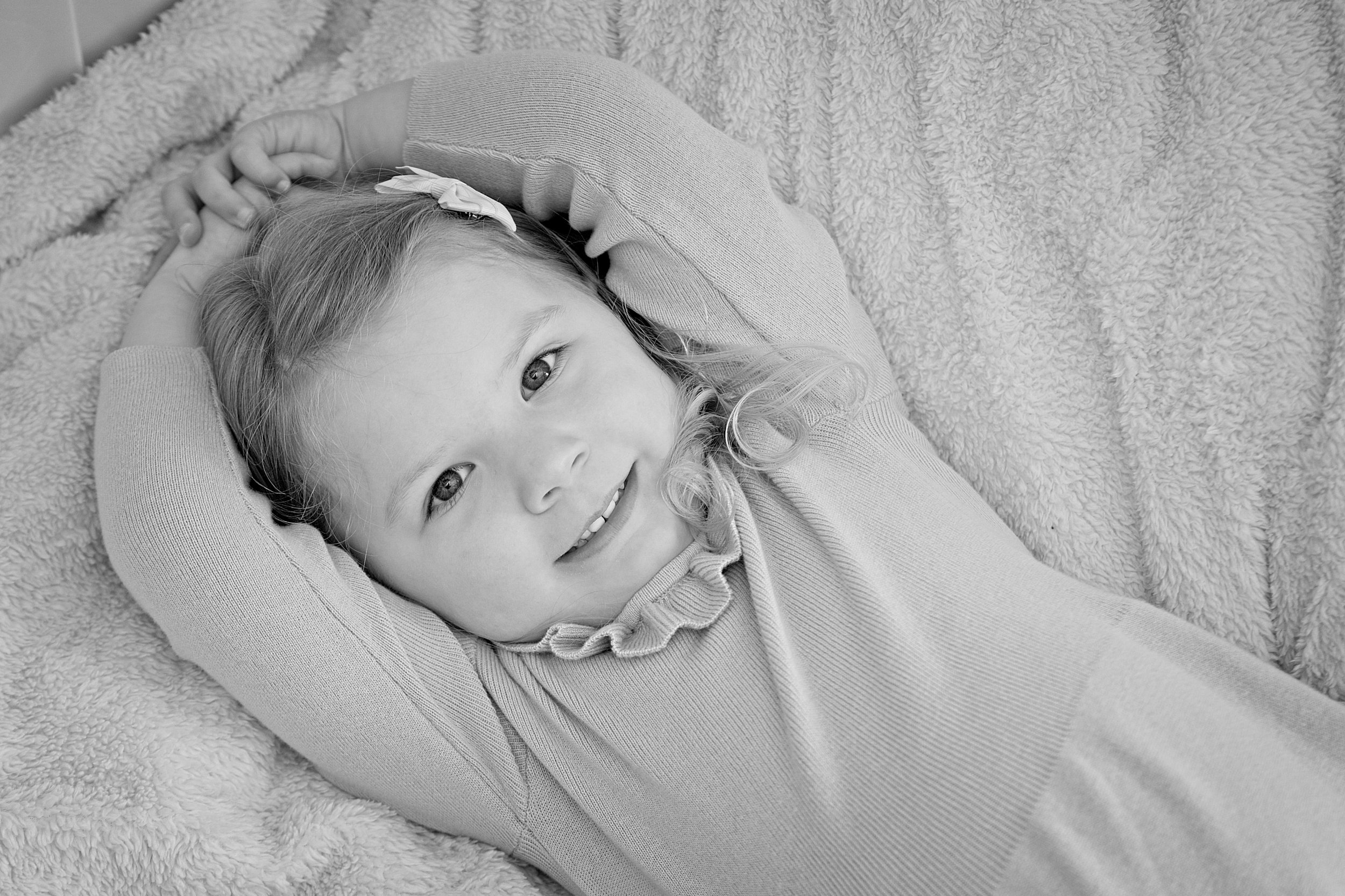 A young girl lying on a plush blanket with her hands behind her head, smiling at the camera, in black and white. Photography by photographer based in Southam, Warwickshire