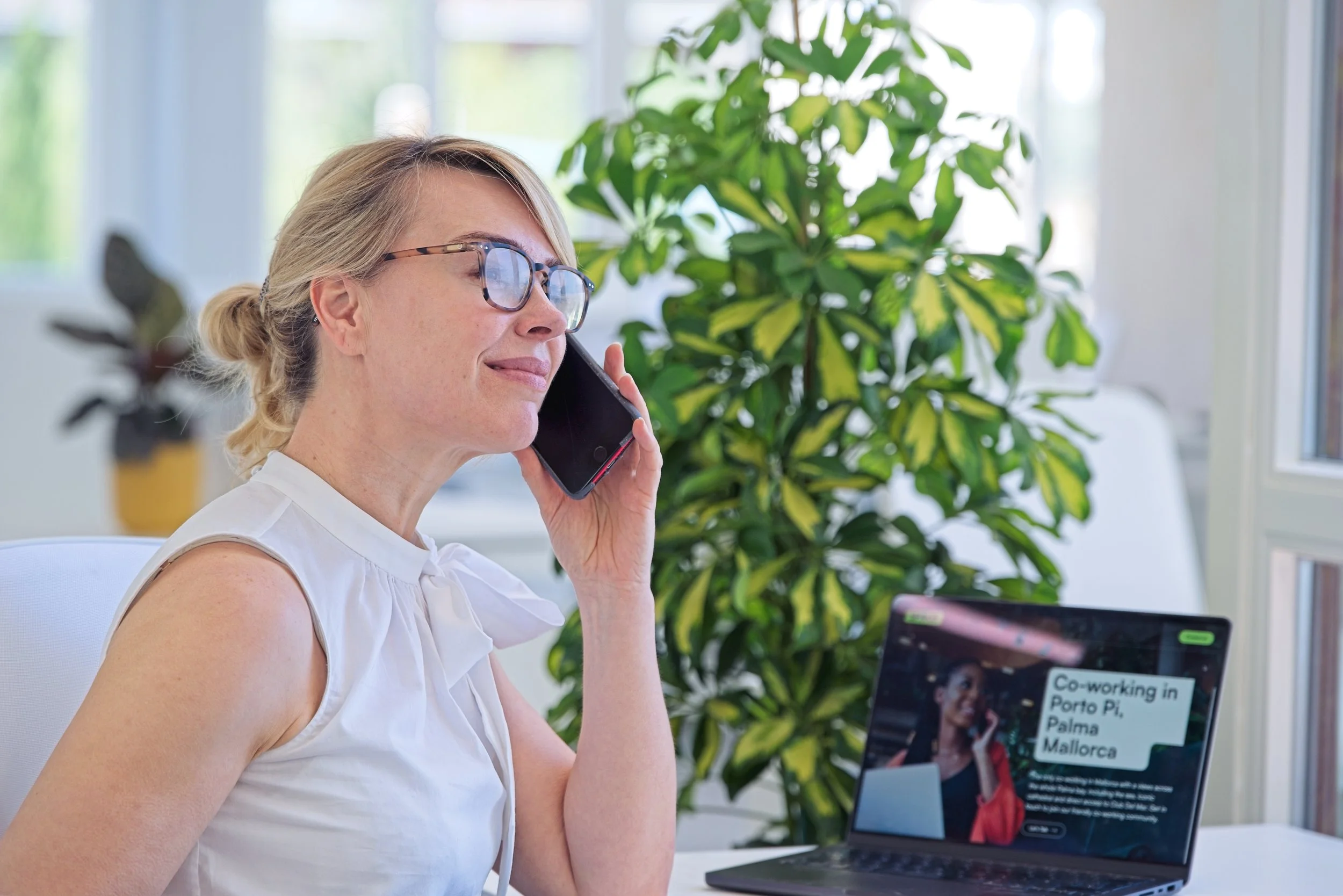 A woman with blonde hair and glasses talking on a cellphone in an office setting. There are large green plants behind her and a laptop on the desk showing a website titled 'Co-working in Porto Pi, Palma Mallorca.'