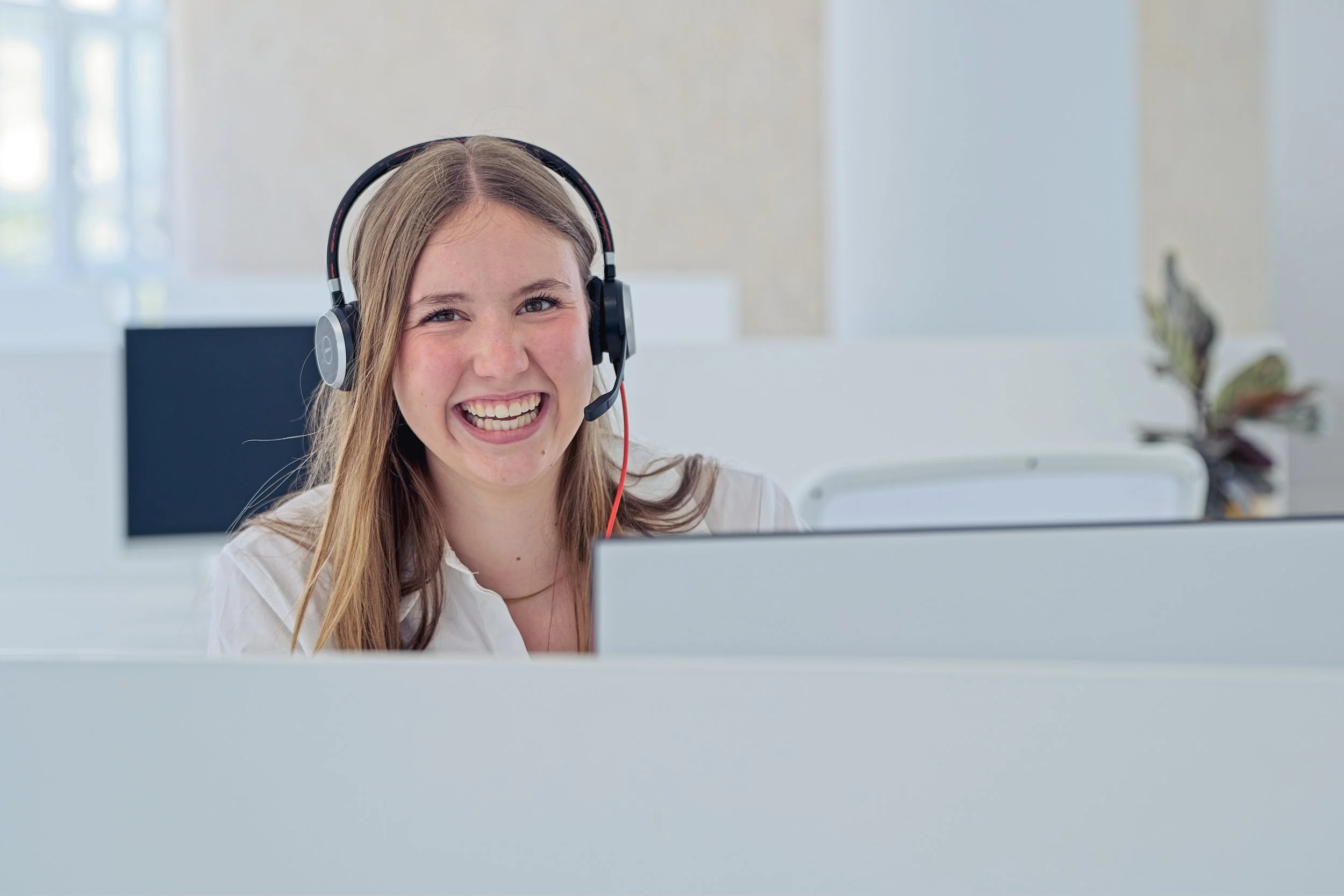 A young woman with long blonde hair smiling and wearing a headset, sitting in front of a computer in a bright office environment.