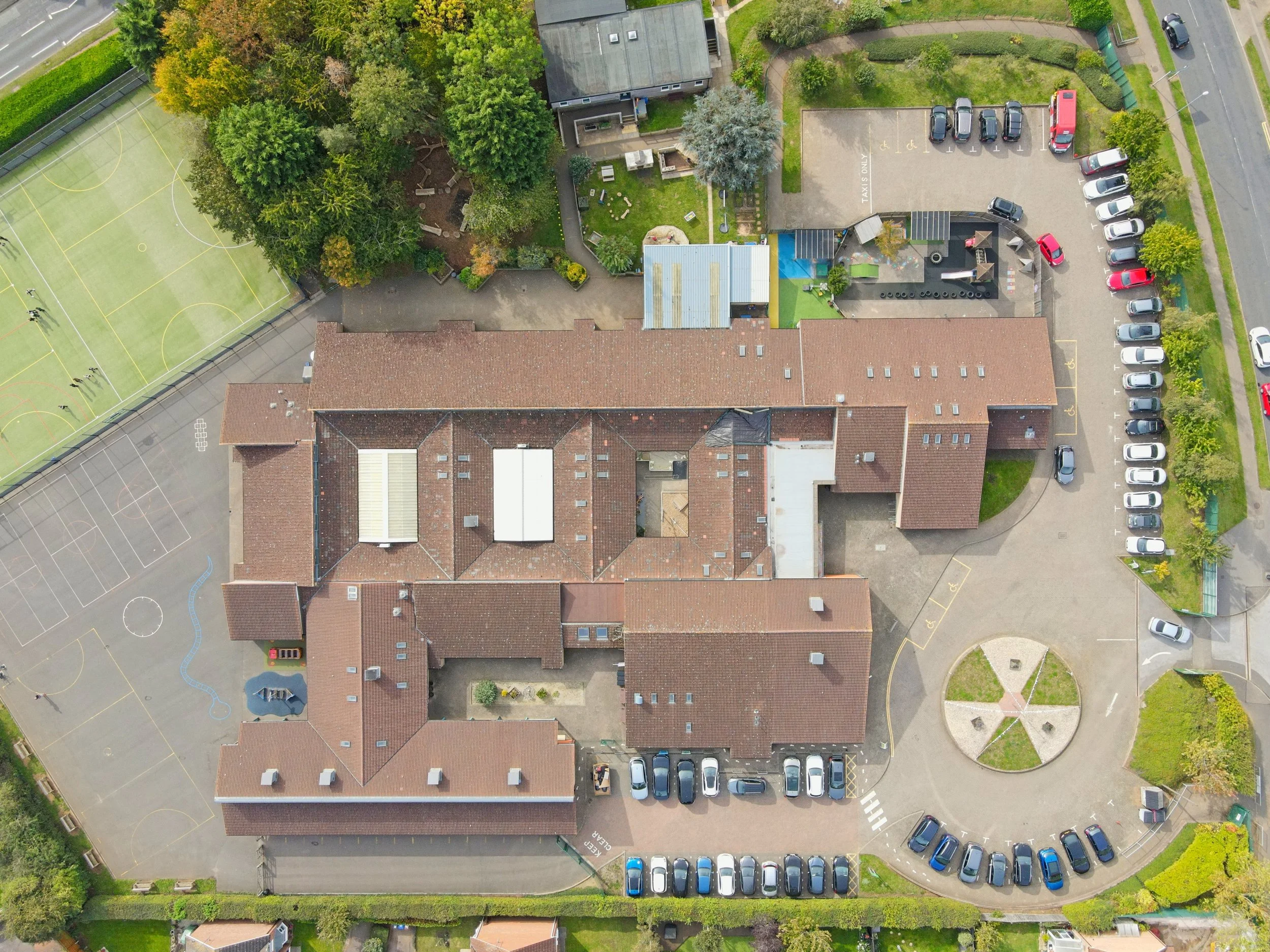 An aerial view of a schoolyard featuring a basketball court, a playground, green trees, parking lot with cars, and surrounding residential houses with red roofs.