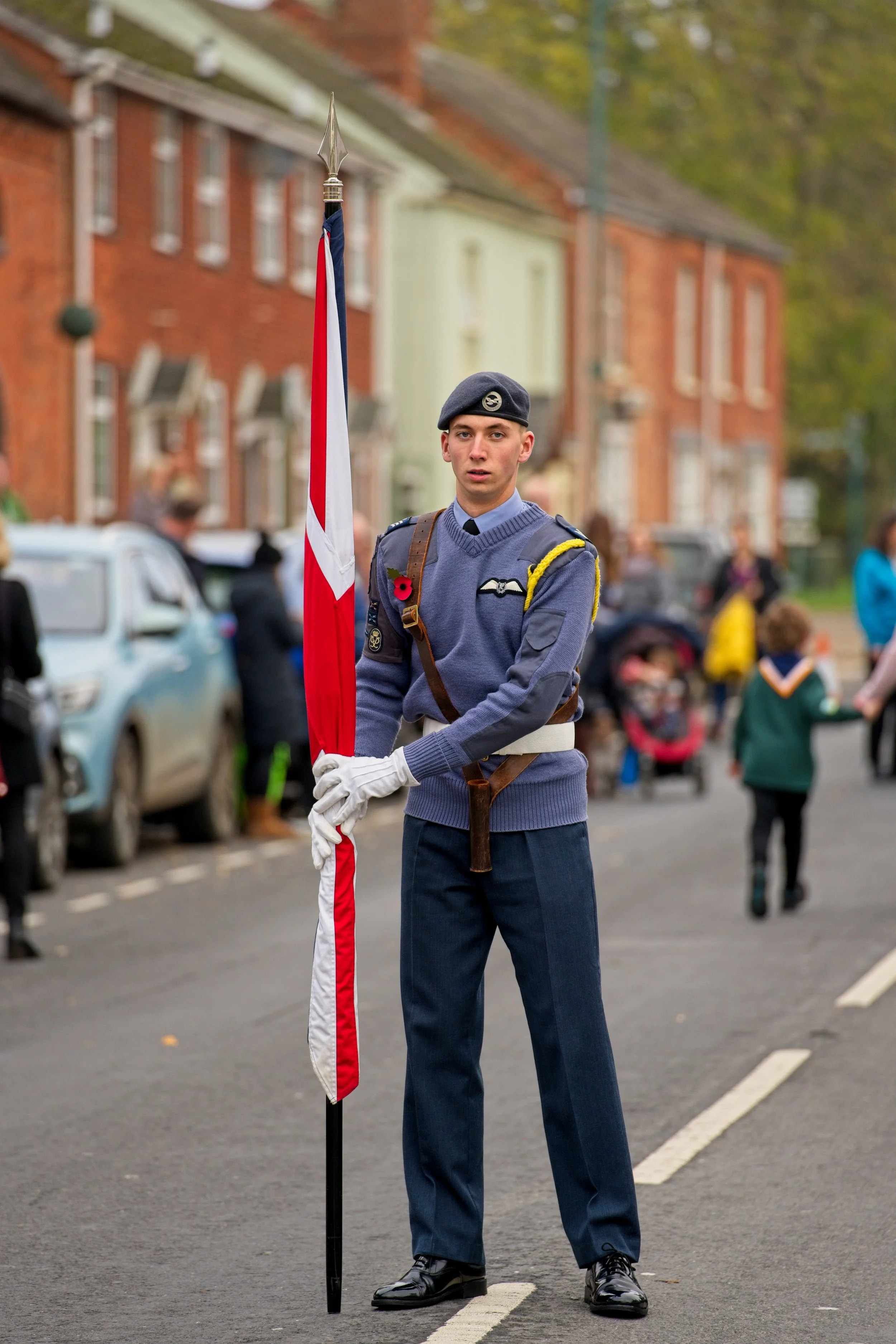 Young man in military uniform holding a flag during a public event on a street with spectators in the background. Photography by photographer based in Southam, Warwickshire