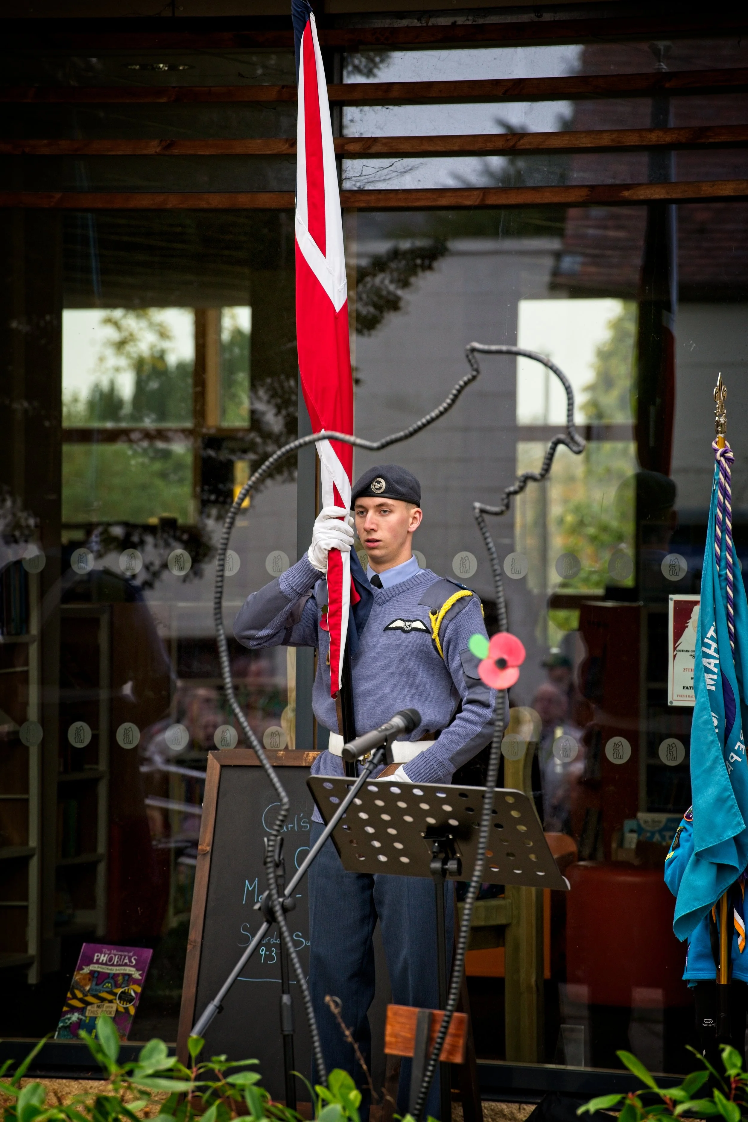 A young man in a military uniform and beret holding a red and white flag, standing behind a microphone and music stand inside a building with glass windows during a memorial or ceremony. Photography by photographer based in Southam, Warwickshire