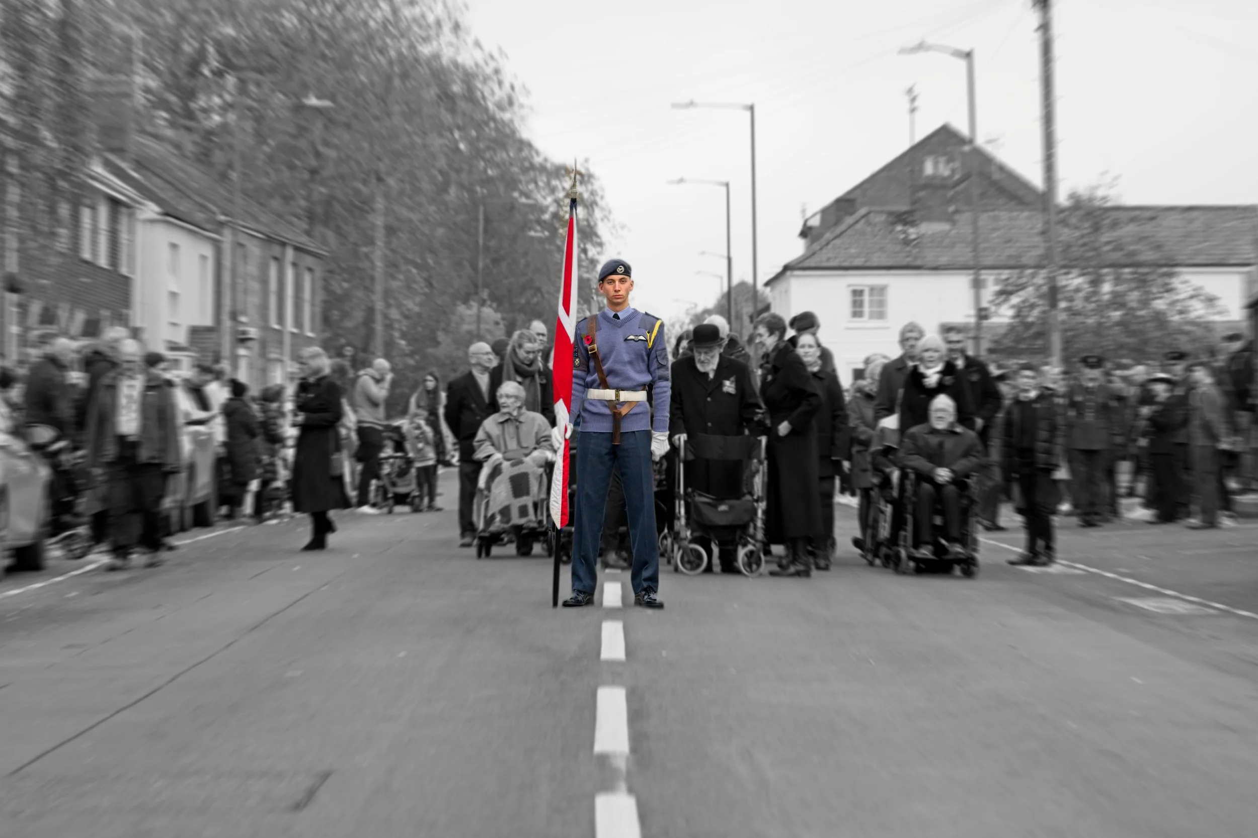 A color image of a soldier standing in the middle of a street holding a flag, with a crowd of people in black and white surrounding him, some with wheelchairs and blankets, and houses in the background.Photography by photographer based in Southam, Wa