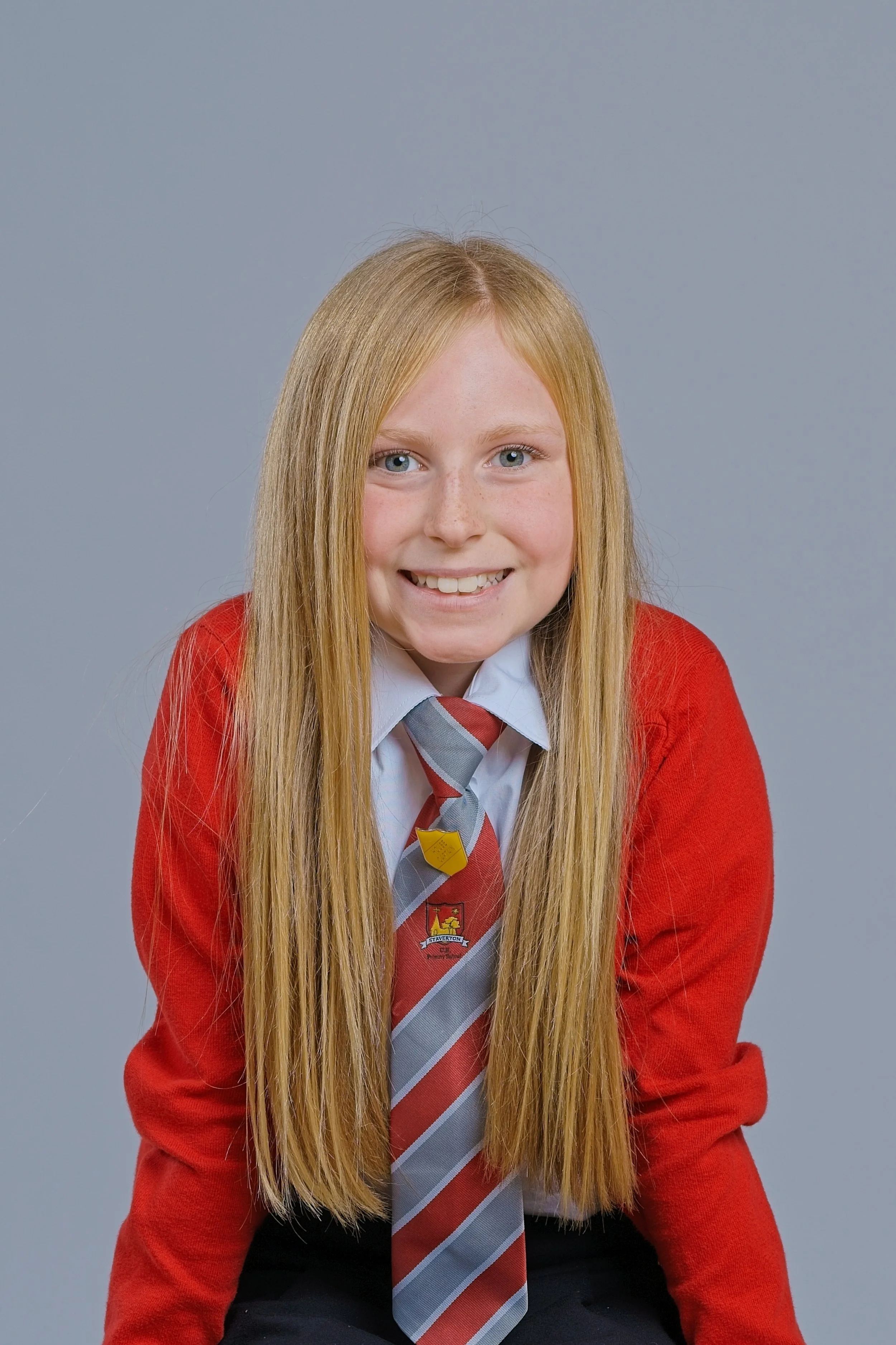 A young girl with long blonde hair wearing a red school sweater, a white shirt, and a gray and red striped tie, smiling at the camera against a gray background.