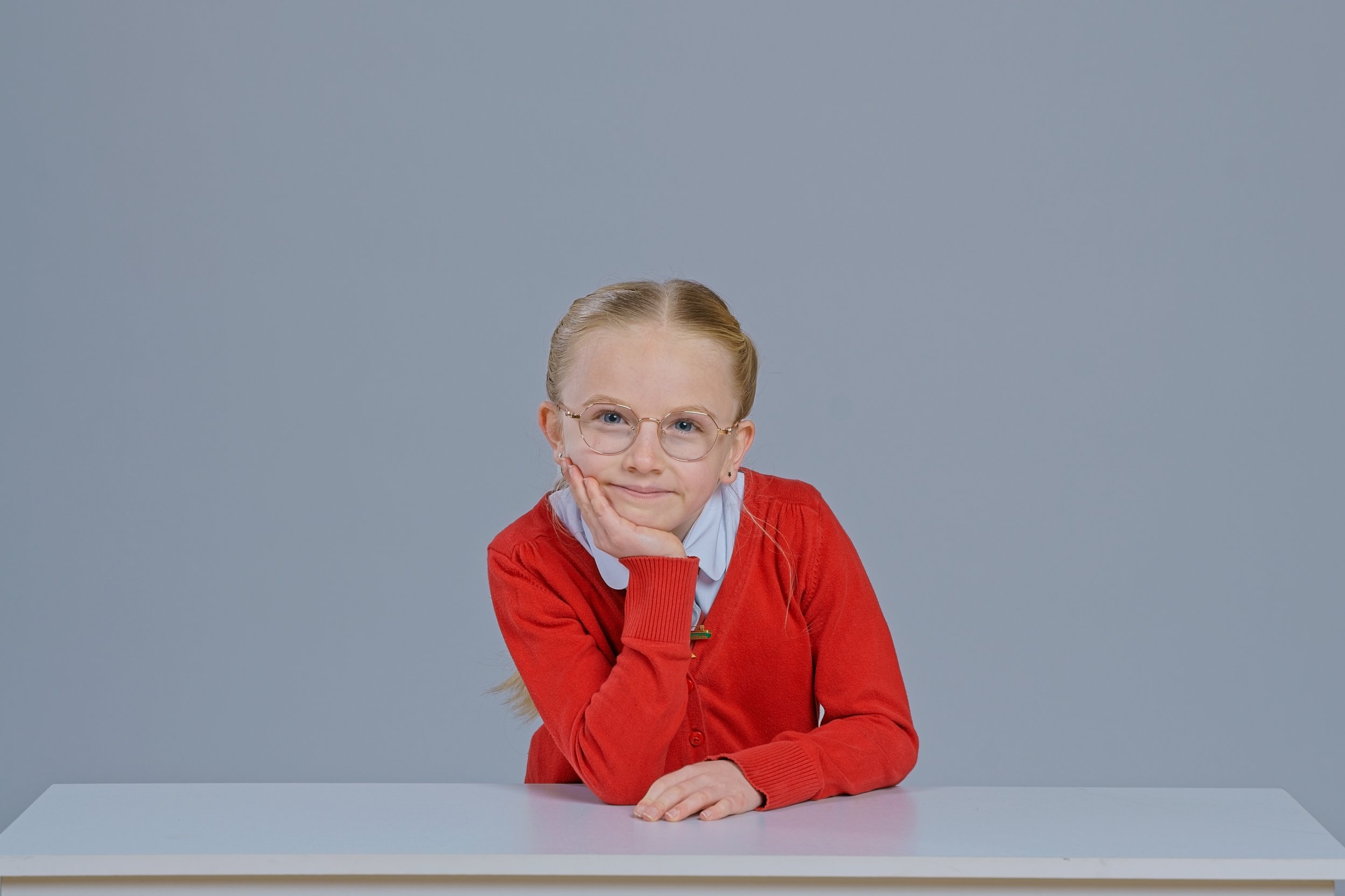 A young girl with blonde hair, glasses, and a red sweater sitting at a table with her chin resting on her hand, against a plain gray background.