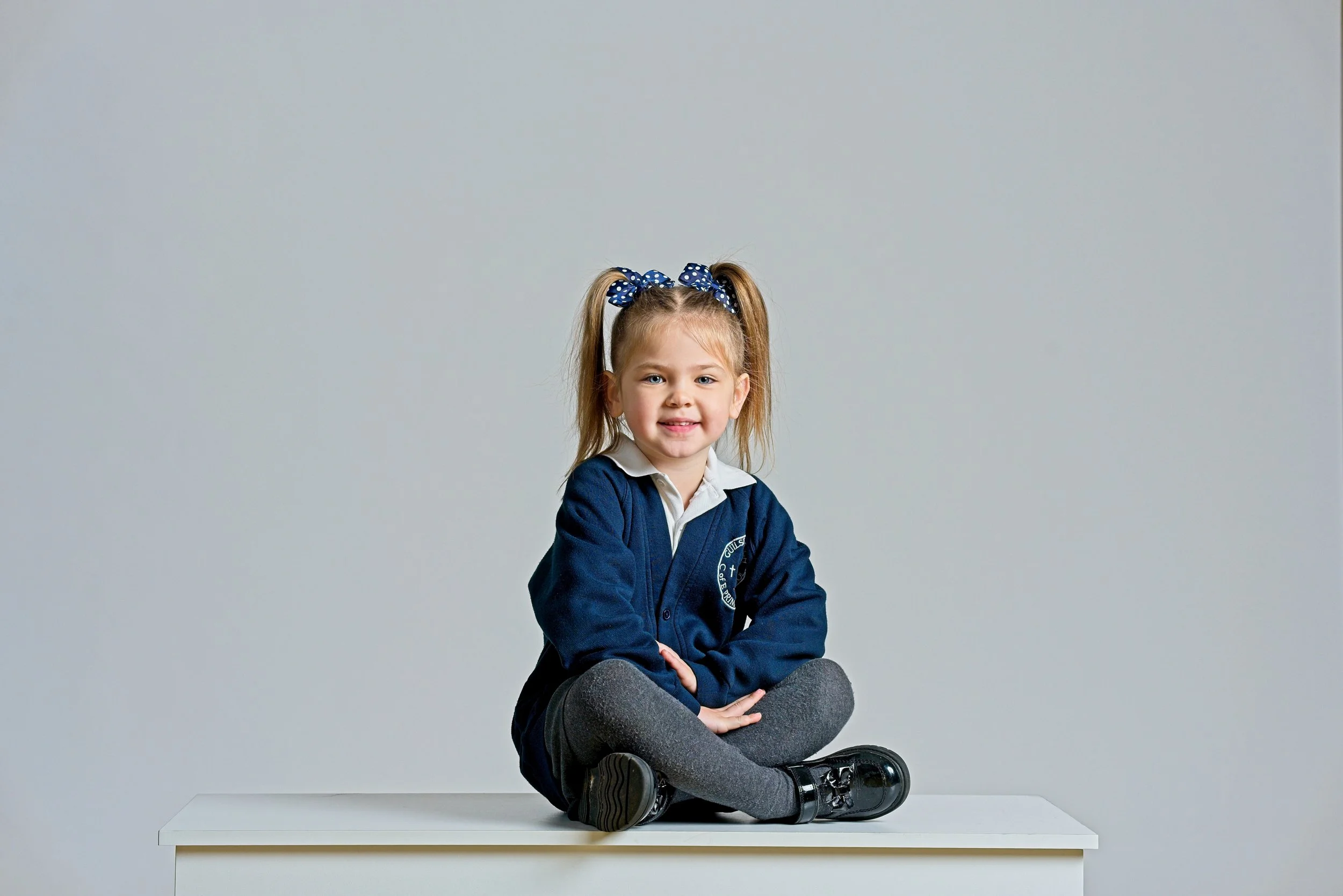 Young girl with blonde hair in pigtails with navy polka-dot bows, wearing a navy sweater with a crest, sitting cross-legged on a white table against a gray background.