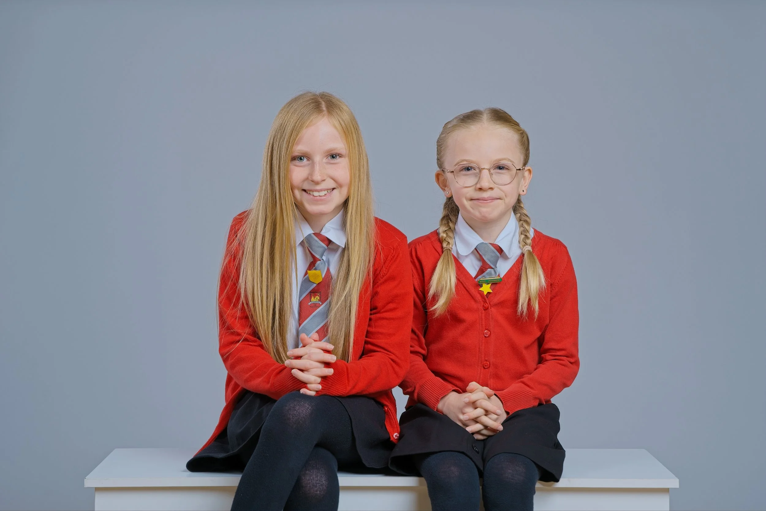 Two young girls in school uniforms with red sweaters and striped ties sitting on a white bench against a plain blue background. One girl has long blonde hair and the other has braided blonde hair and wears glasses.