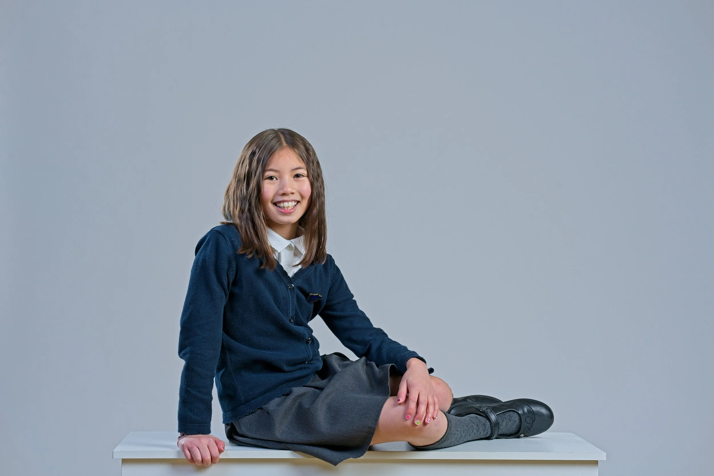 A young girl with long brown hair, smiling, sitting on a white table with one knee up, wearing a navy blue sweater over a white shirt, gray skirt, gray socks, and black shoes, against a plain gray background.
