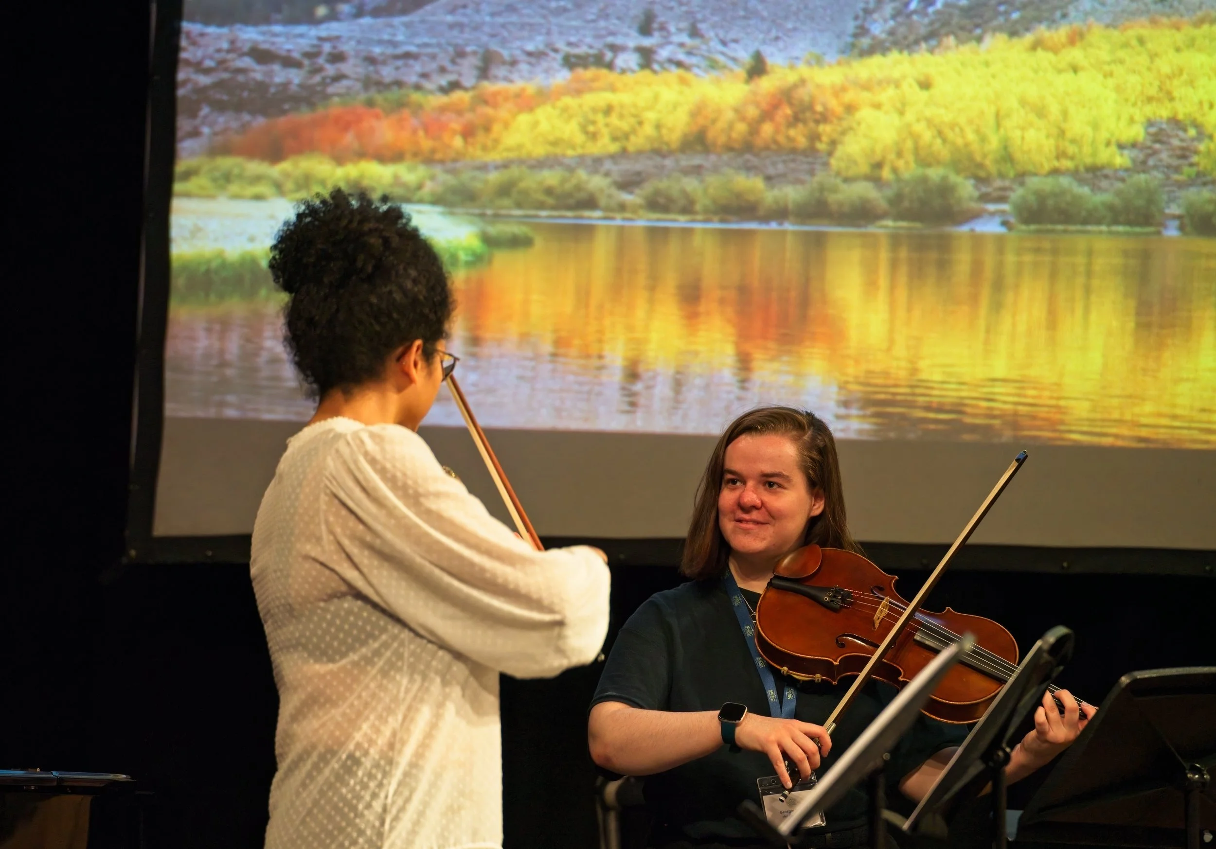Two young women with musical instruments, a violin and a viola, perform together in front of a screen displaying a scenic landscape of a lake and trees with fall foliage. Event Photography by photographer based in Southam, Warwickshire