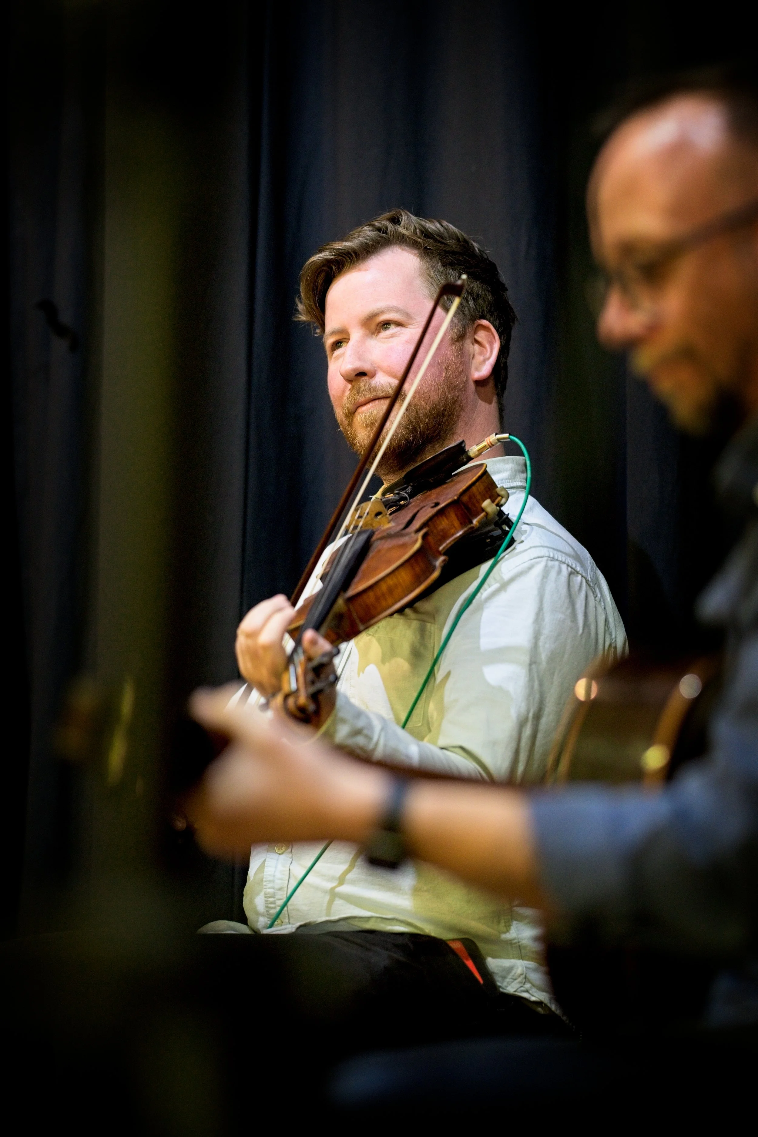 Two musicians playing string instruments, with a man in focus holding a violin and a man on the side playing guitar, during a performance. Event Photography by photographer based in Southam, Warwickshire
