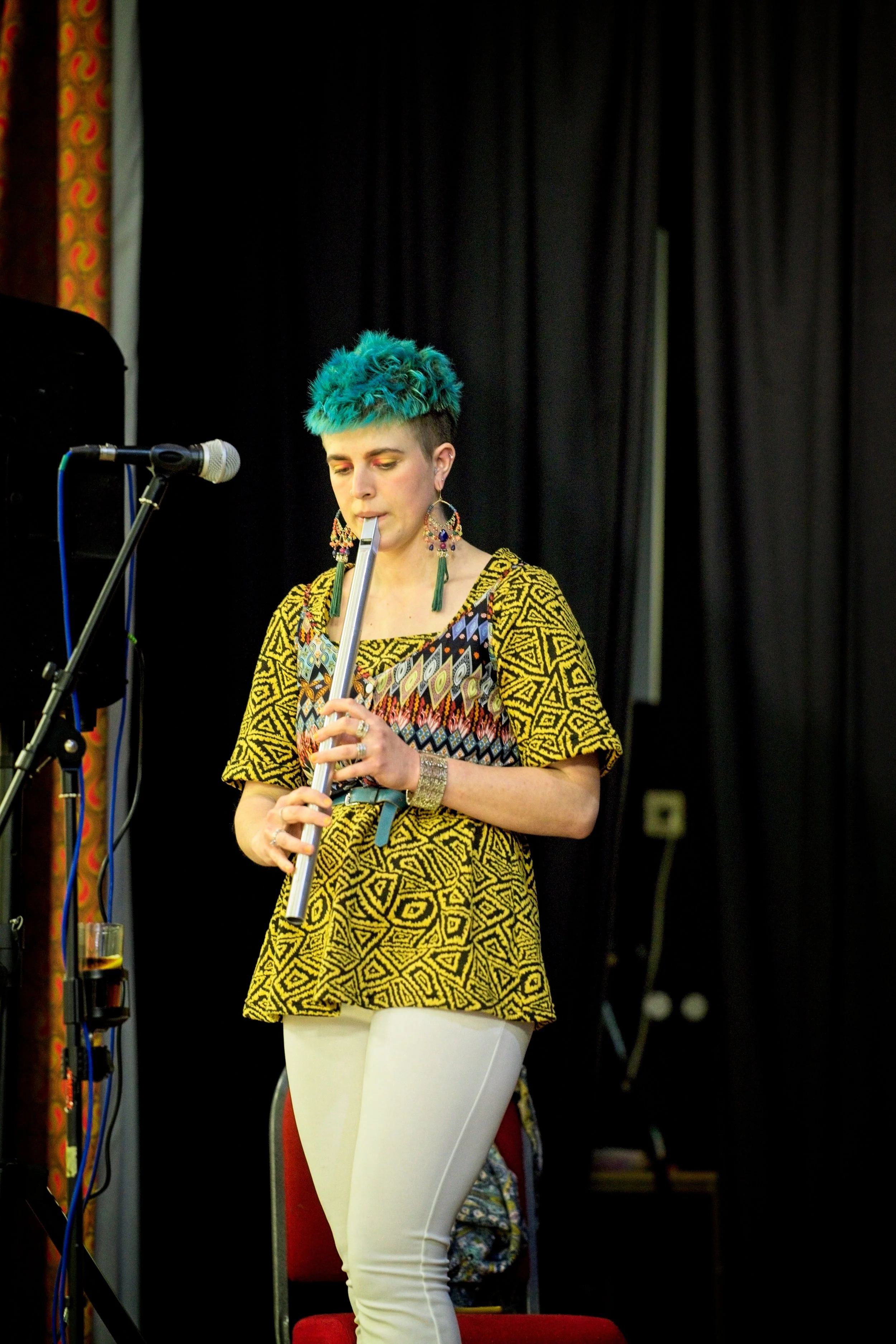 A woman with bright blue hair playing a silver flute on stage, wearing a yellow patterned top and white pants, with a microphone stand nearby. Event Photography by photographer based in Southam, Warwickshire