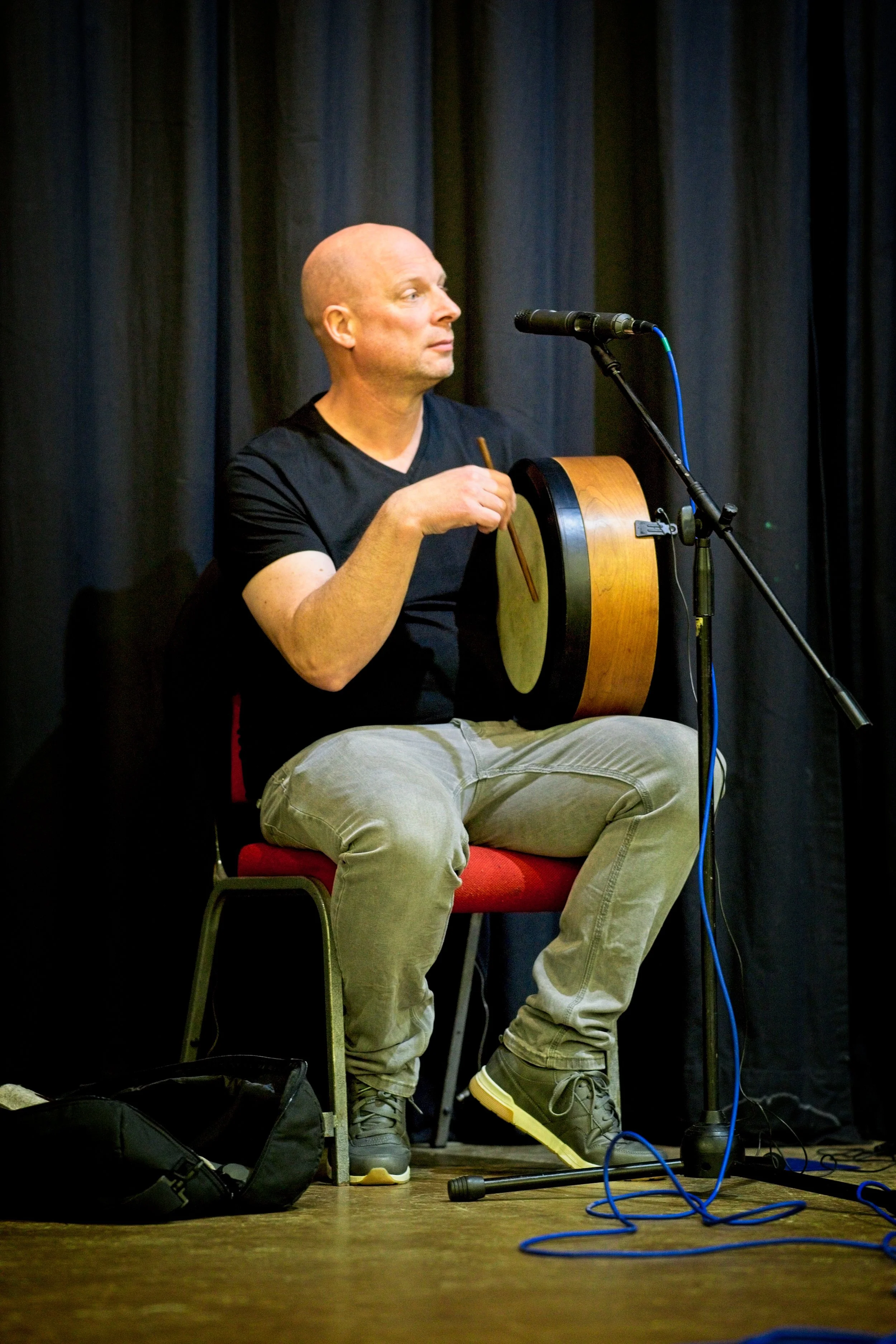 A man with a shaved head and black t-shirt sitting on a red chair, playing a drum with a drumstick, positioned next to a microphone on a stage with black curtains in the background. Event Photography by photographer based in Southam, Warwickshire