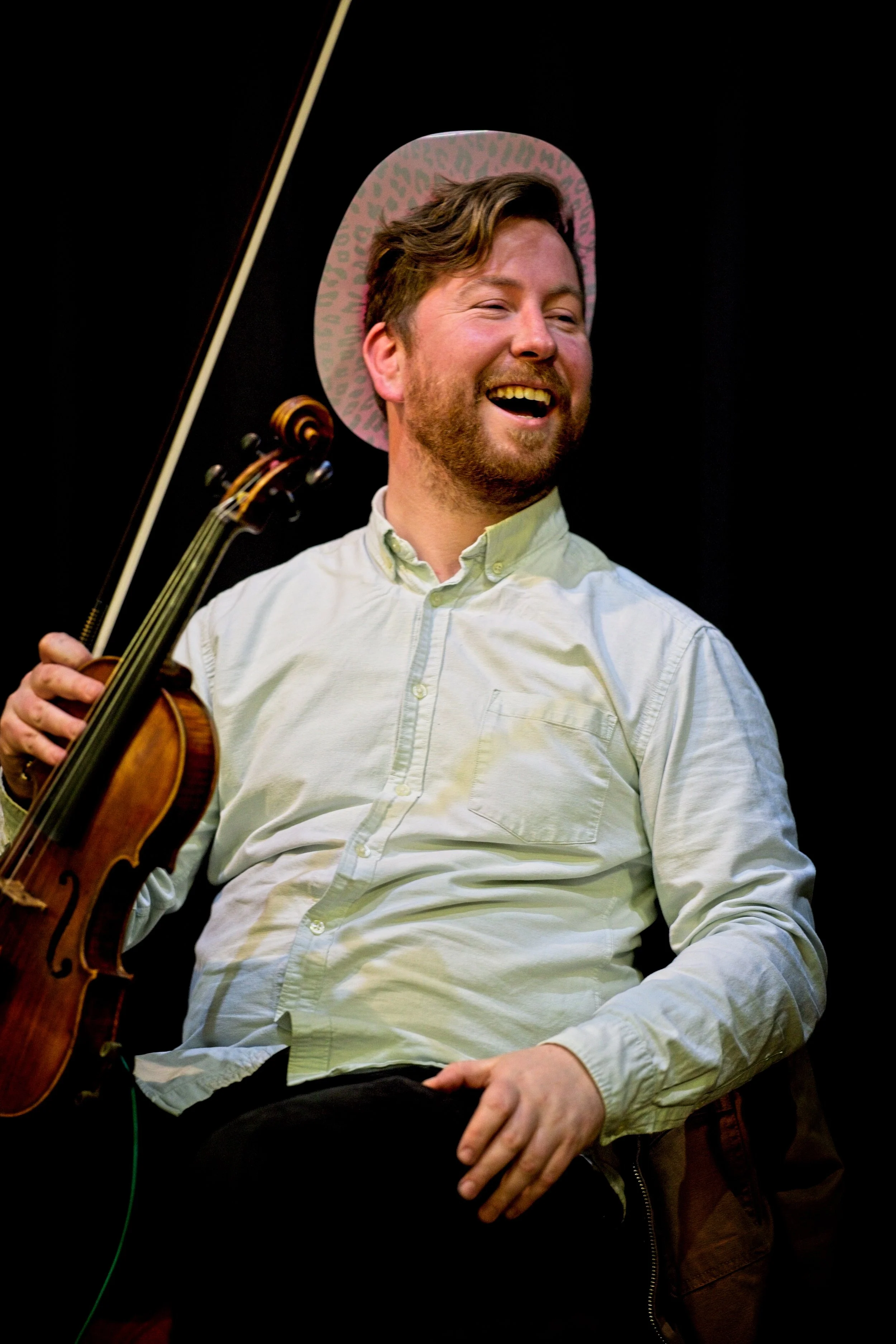 A man with a beard laughing, holding a violin, wearing a pink leopard print hat, and sitting against a dark background. Event Photography by photographer based in Southam, Warwickshire