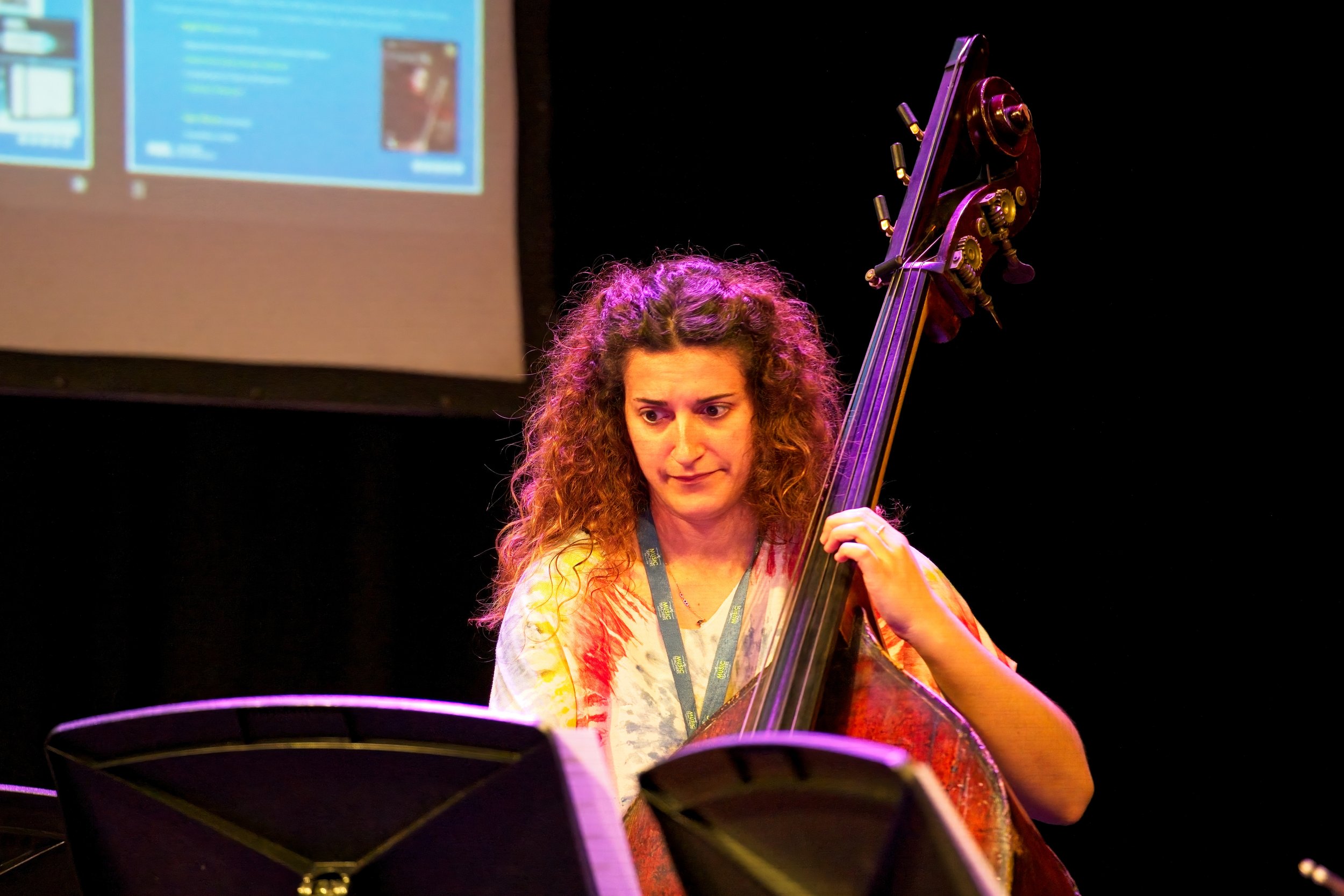 Woman with curly hair playing a double bass on stage with a music stand and a screen in the background. Event Photography by photographer based in Southam, Warwickshire