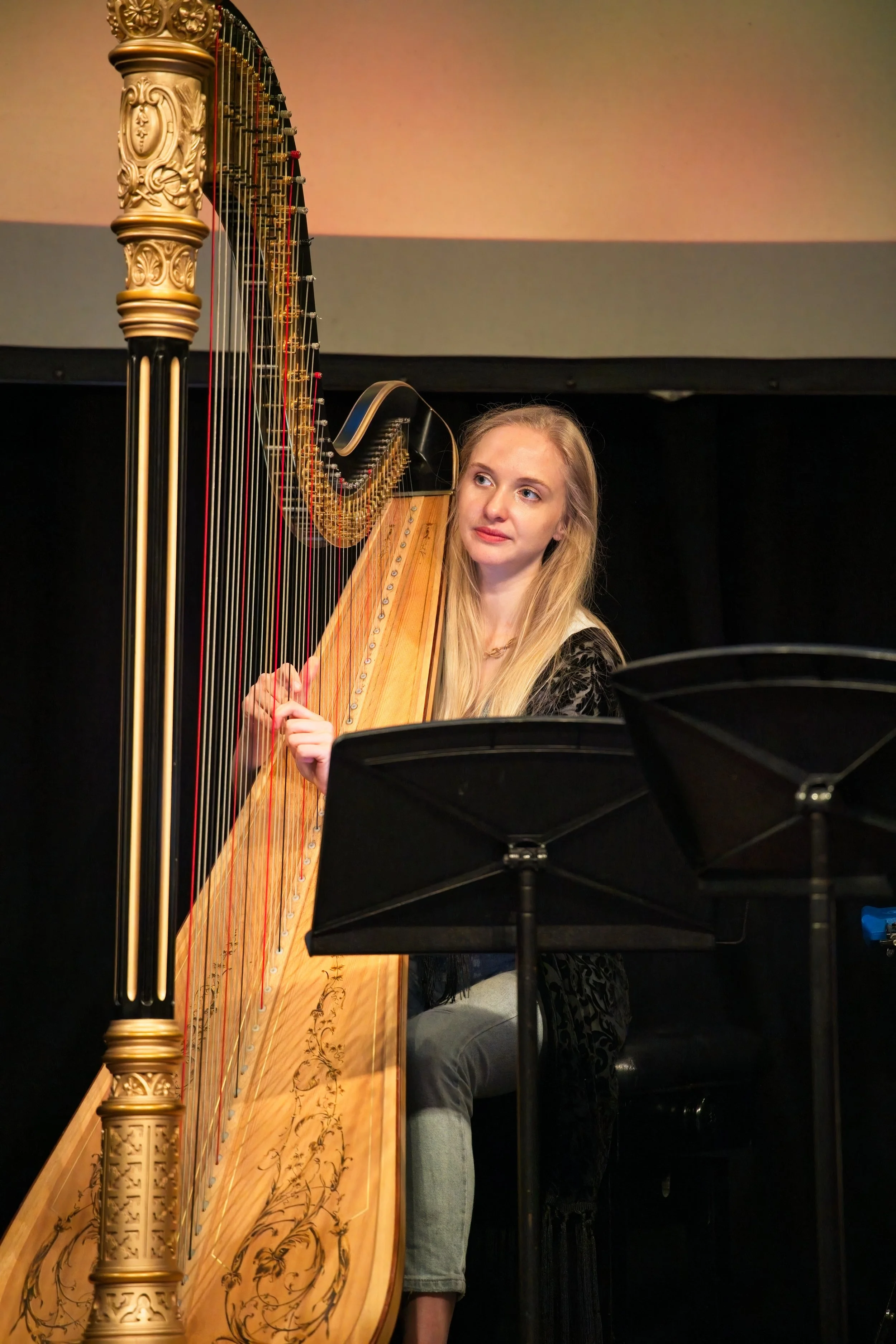 A young woman with long blonde hair playing a large, ornate harp in a dark room, sitting on a black bench with music stands nearby. Event Photography by photographer based in Southam, Warwickshire
