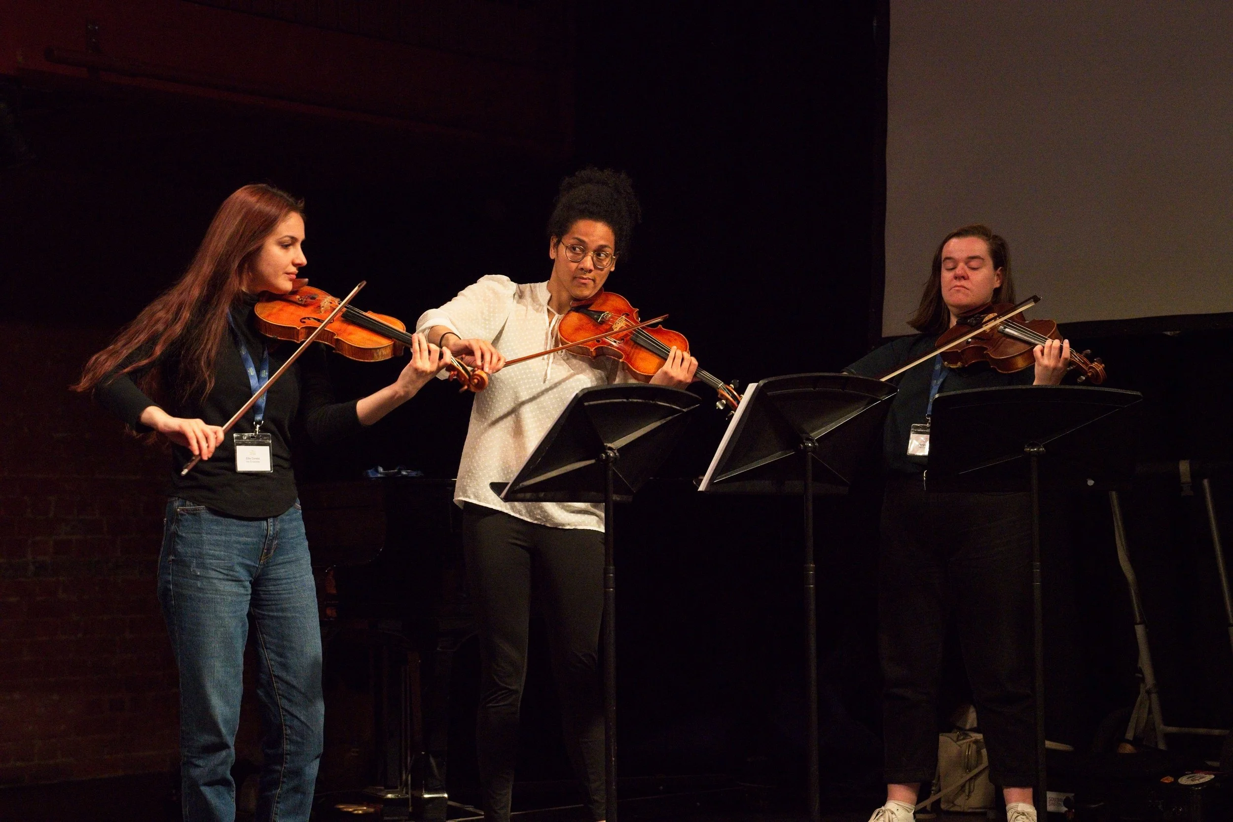 Three women playing violins on stage during a performance, with music stands and a dark background. Event Photography by photographer based in Southam, Warwickshire