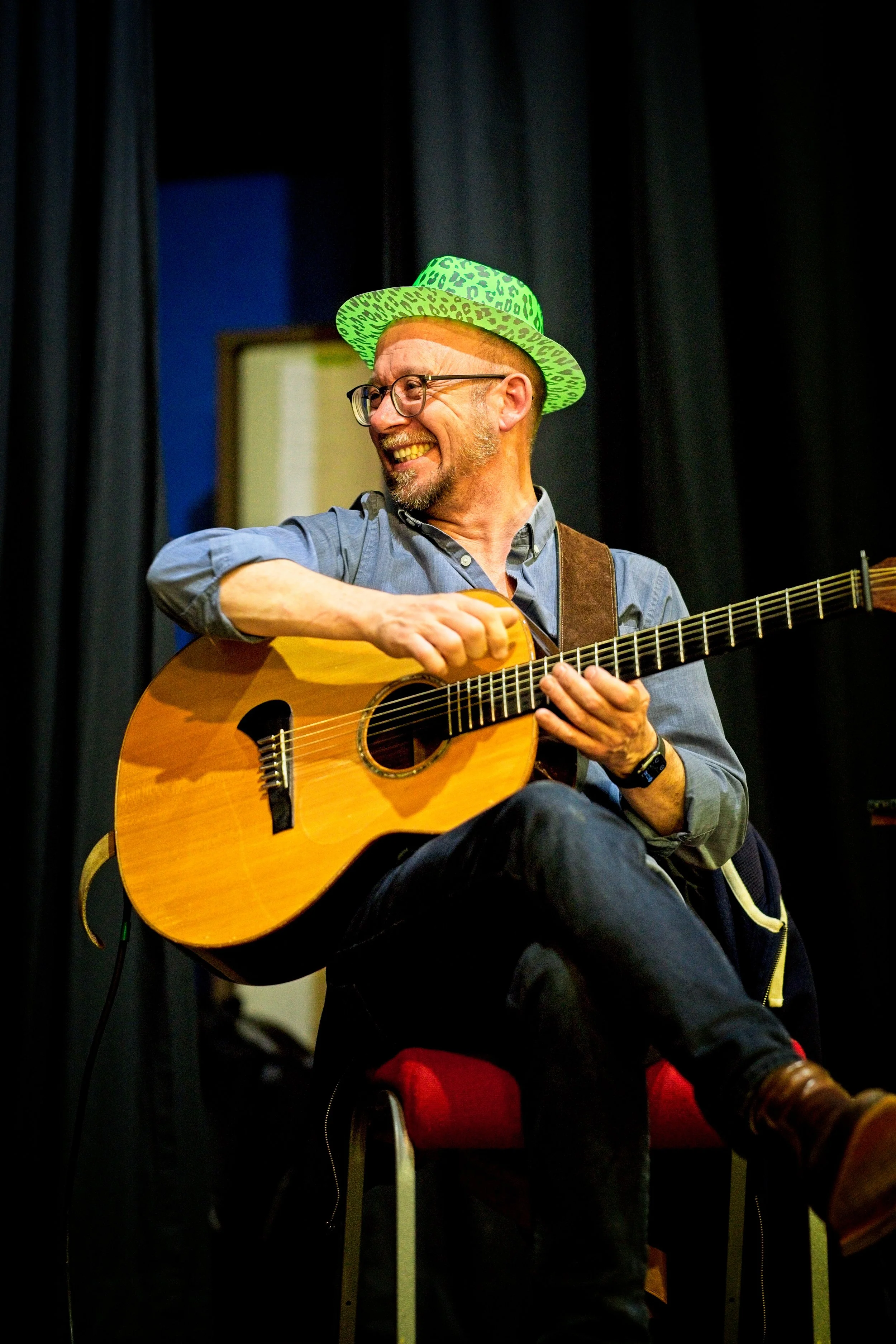 A man with glasses, a beard, and a green leopard-print hat sitting on a red chair, playing an acoustic guitar and smiling. Event Photography by photographer based in Southam, Warwickshire