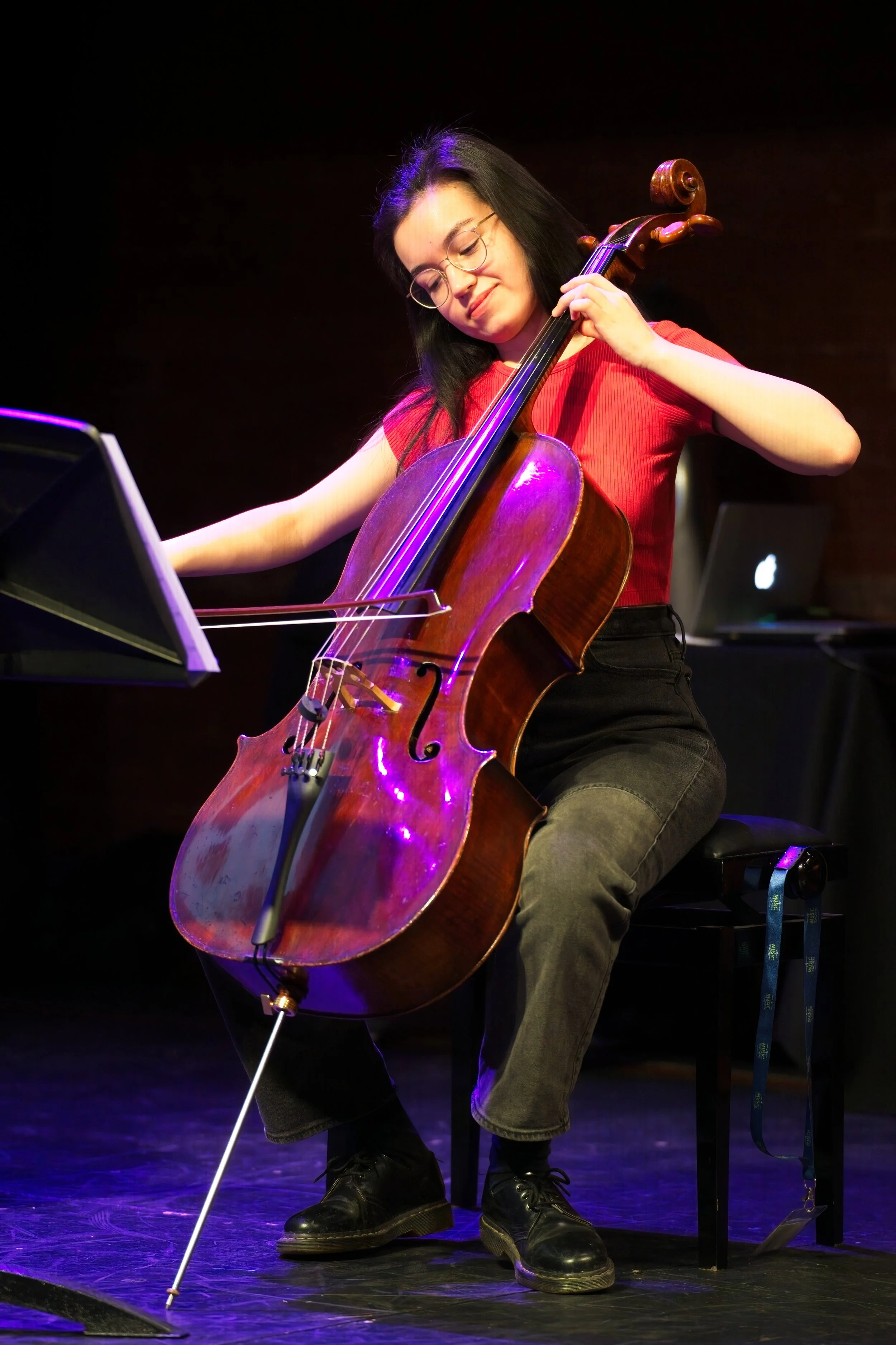 A woman playing a cello during a live performance on stage, lit with purple and yellow lighting, with music stand and laptop nearby. Event Photography by photographer based in Southam, Warwickshire