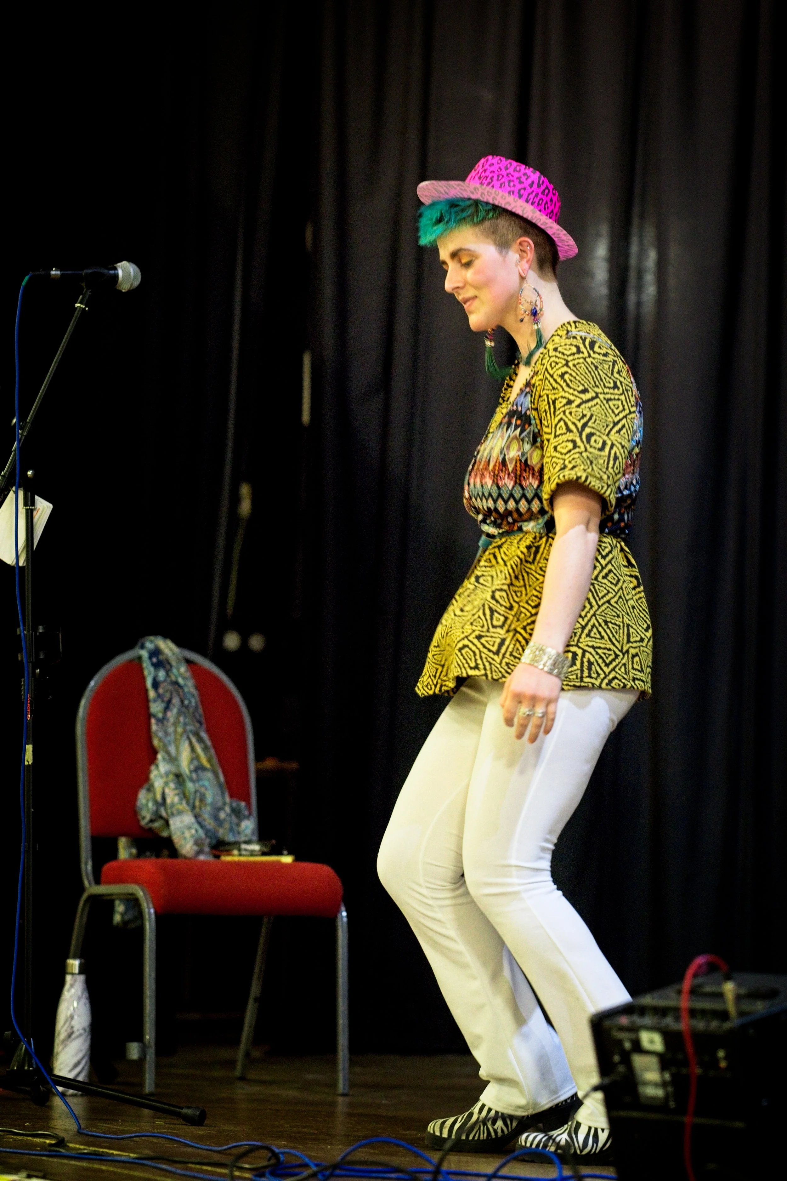 A woman with short green hair, wearing a pink leopard print hat, a colorful embroidered top, a yellow patterned blazer, white pants, and zebra-striped shoes, is dancing on a stage with black curtains. Event Photography by photographer based in Southa