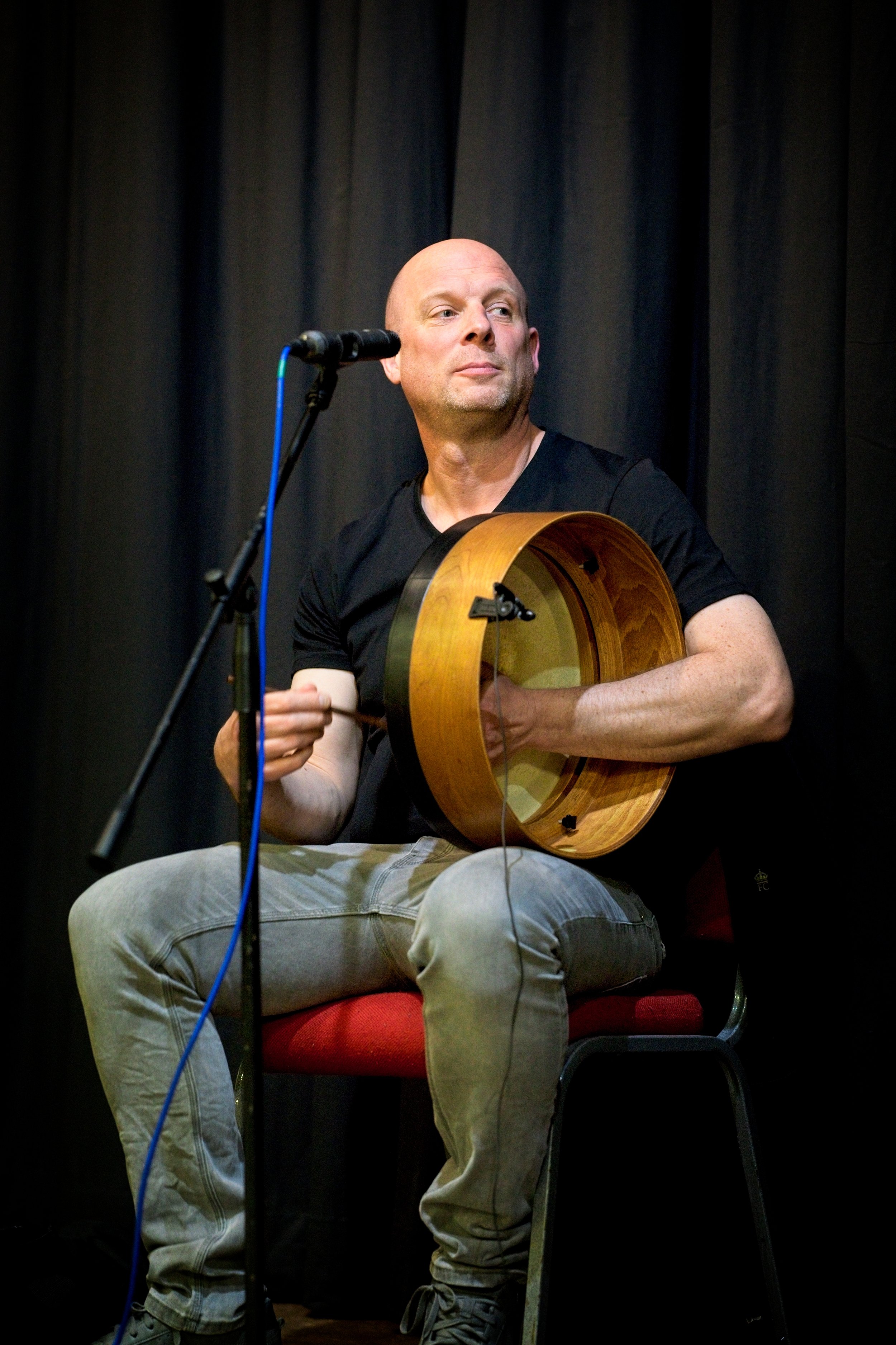 A man sitting on a red chair on stage, holding a Taylor guitar with no strings, set against black curtains, while looking to his right. Event Photography by photographer based in Southam, Warwickshire