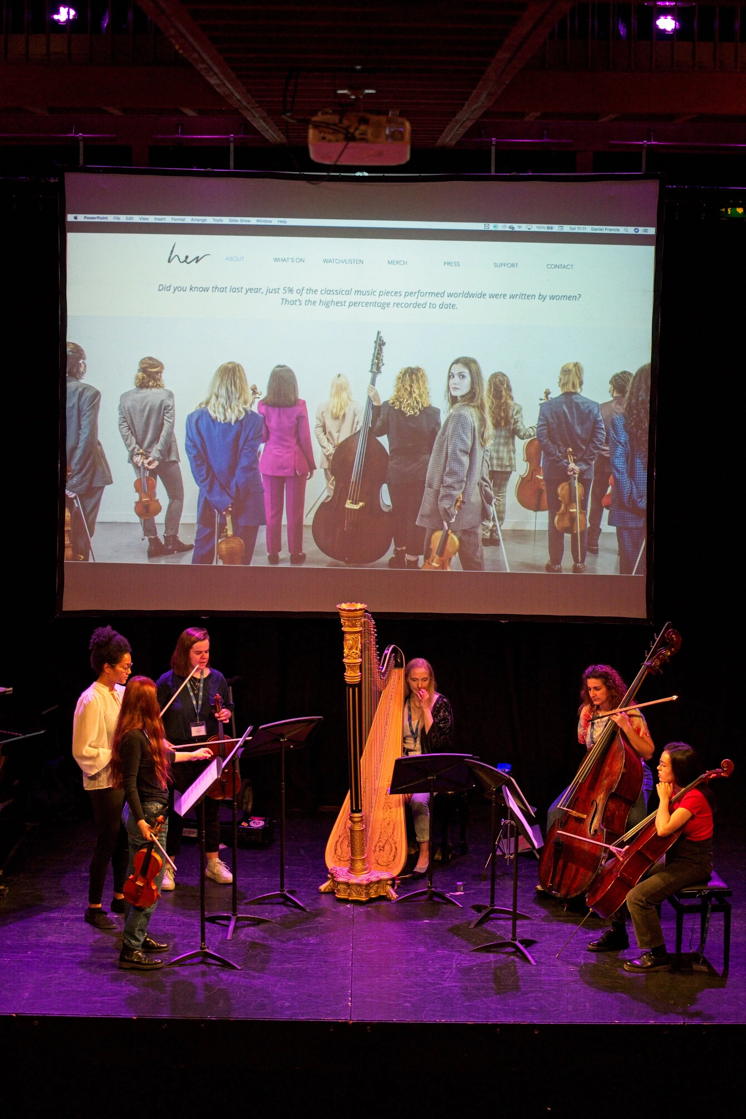 A group of women musicians on stage with a harp and string instruments, performing in front of a large screen displaying a website with women holding string instruments, and text about women in classical music. Event Photography Warwickshire