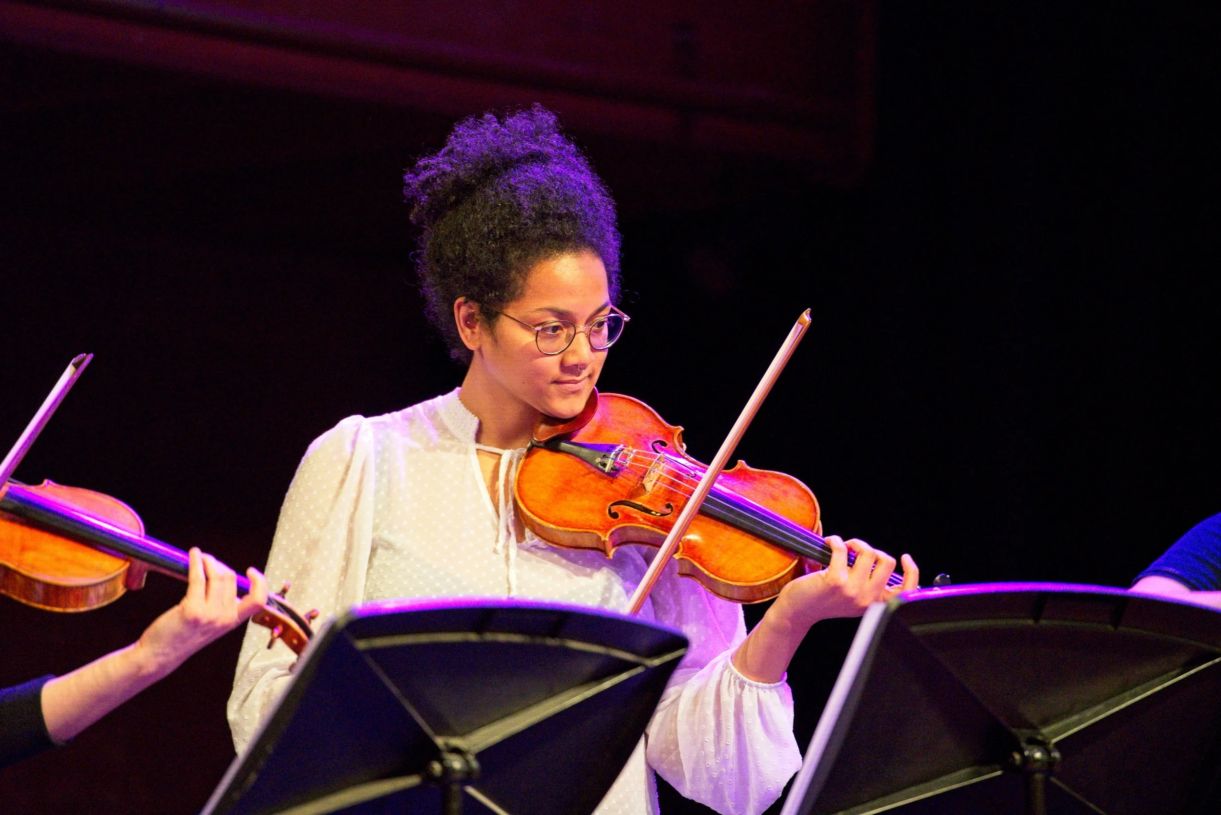 A woman with glasses and curly hair playing a violin during a performance, with music stands in front of her, dark background, and purple stage lighting. Event Photography by photographer based in Southam, Warwickshire