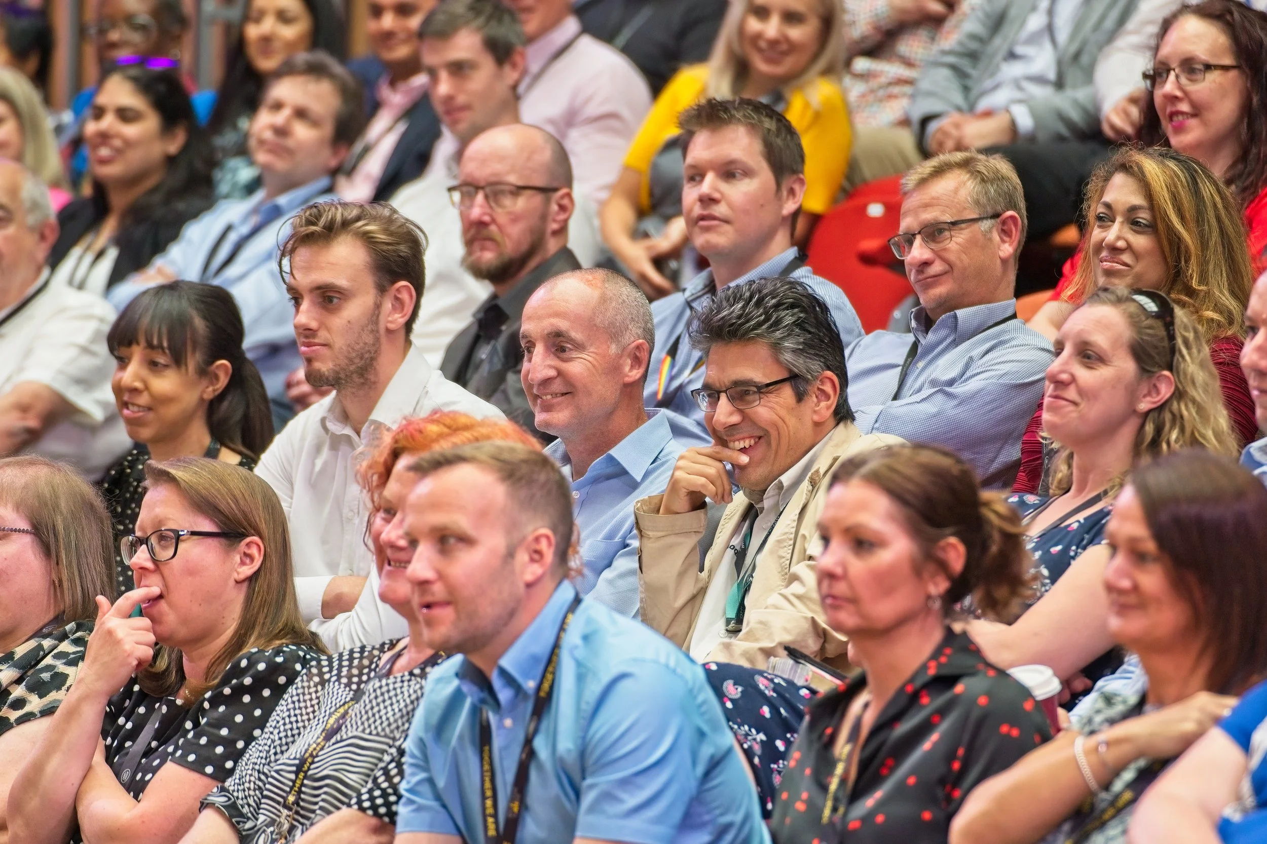 A diverse group of people sitting in an audience at a conference or seminar, attentively listening and smiling. Event Photography by photographer based in Southam, Warwickshire.