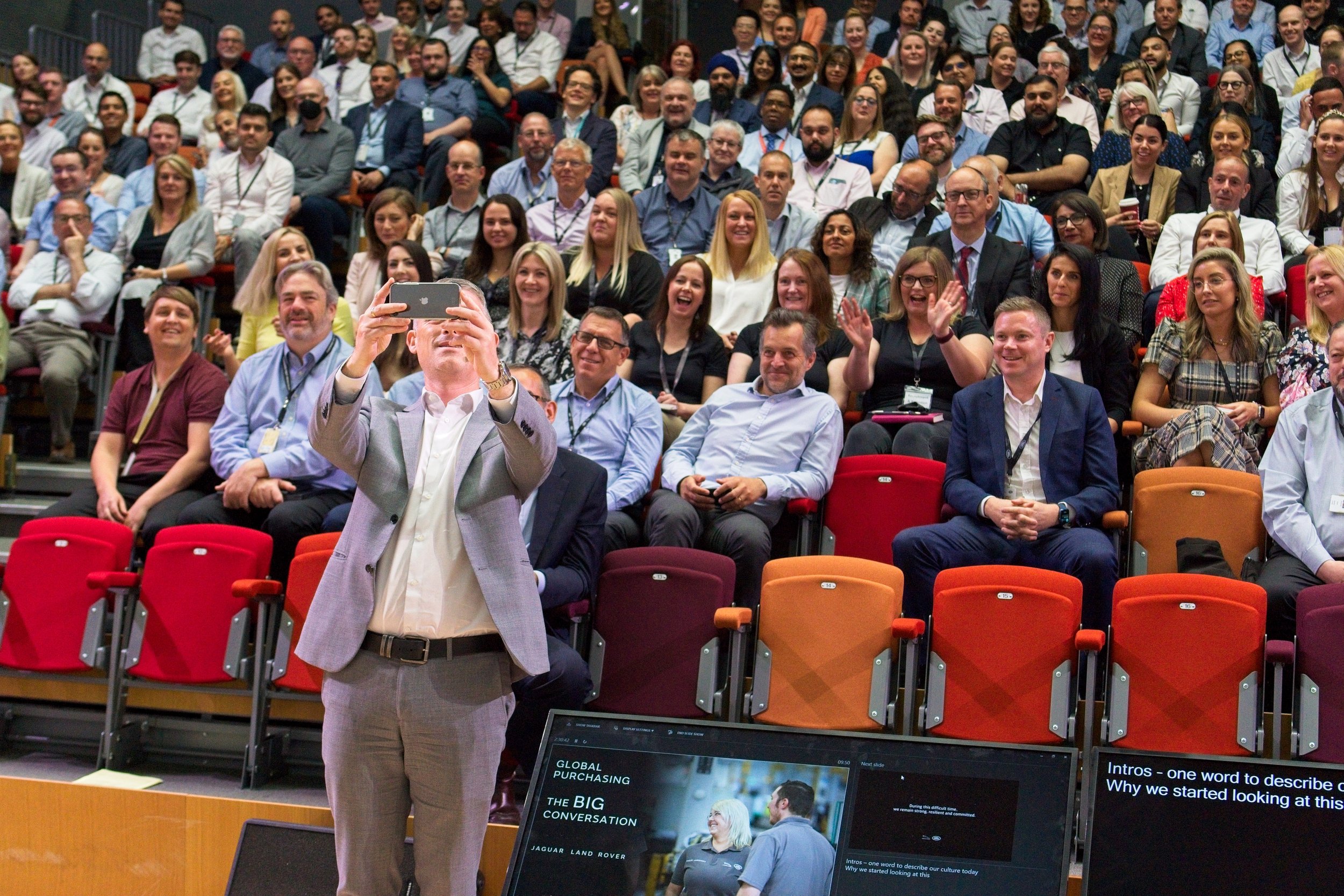 Man in light gray suit taking a picture with a smartphone during a conference with a large audience seated in tiered seats. Event Photography by photographer based in Southam, Warwickshire.