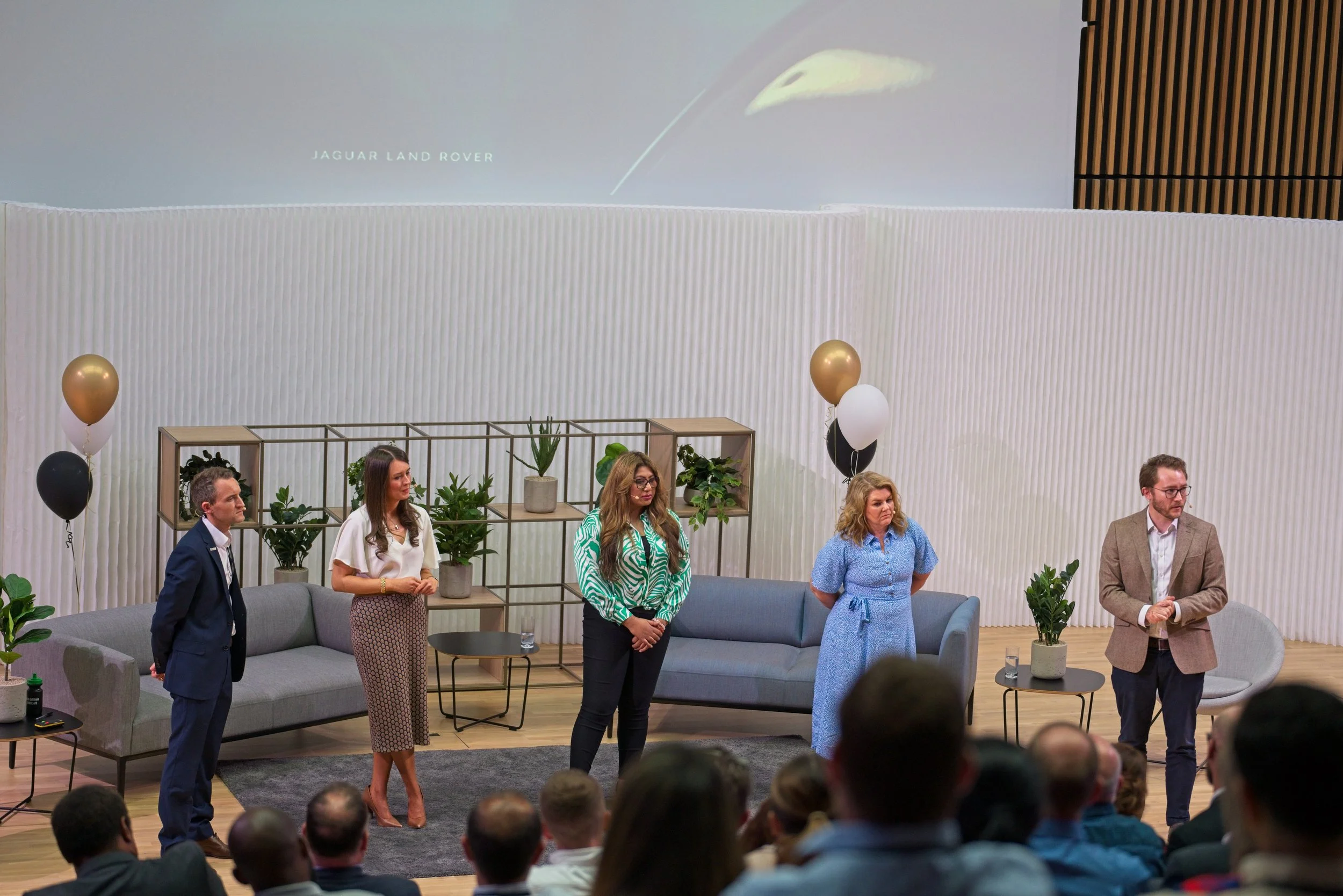 Five people standing on a stage with a seated audience in front, behind them is a white wall, potted plants, and a large screen displaying a leaf and the text 'Jaguar Land Rover'. Event Photography by photographer based in Southam, Warwickshire.
