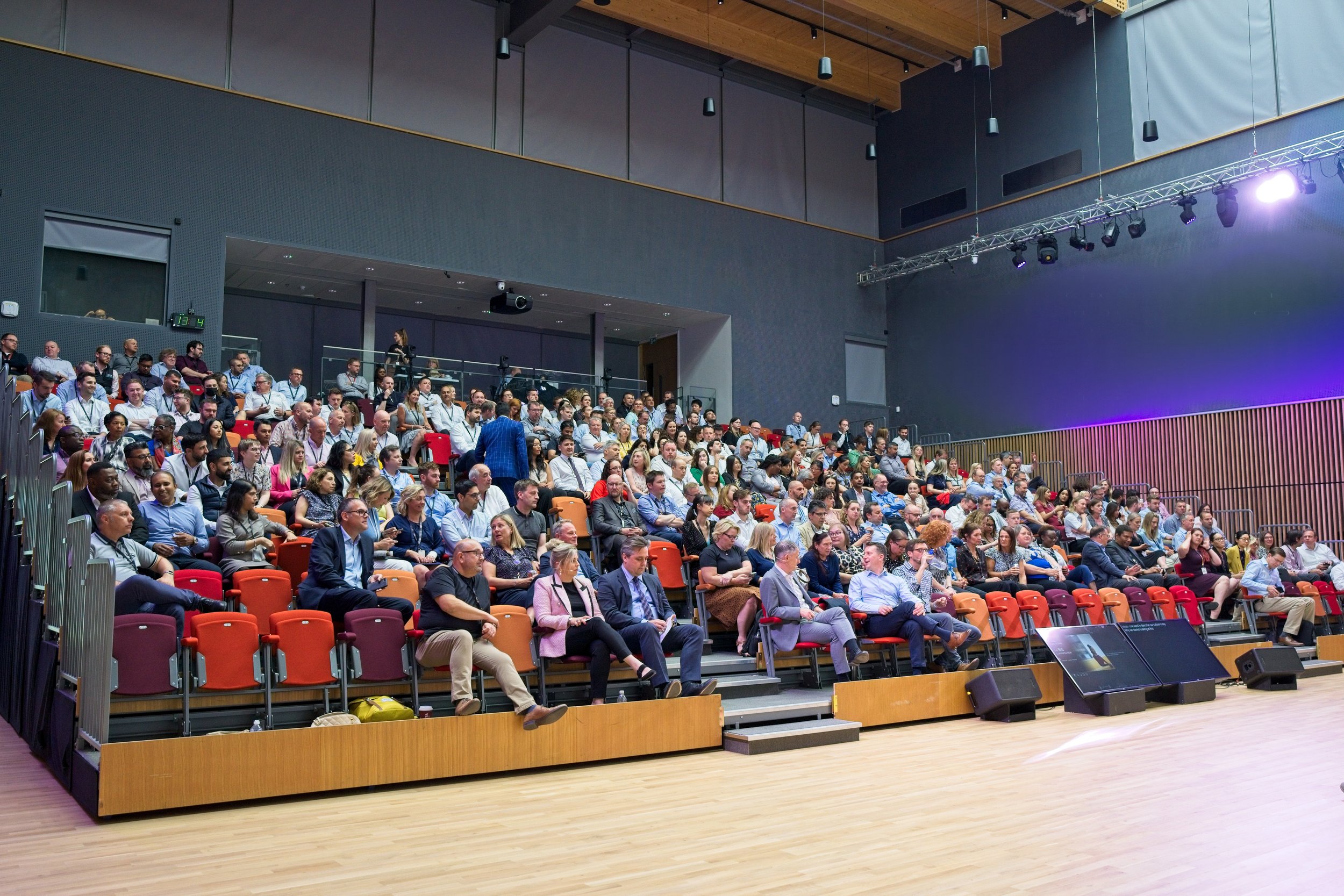 An audience seated in an auditorium, watching a presentation or event on stage. The auditorium has multiple rows of chairs with many attendees, some using electronic devices. The stage has screens and lighting equipment. Event Photography Southam