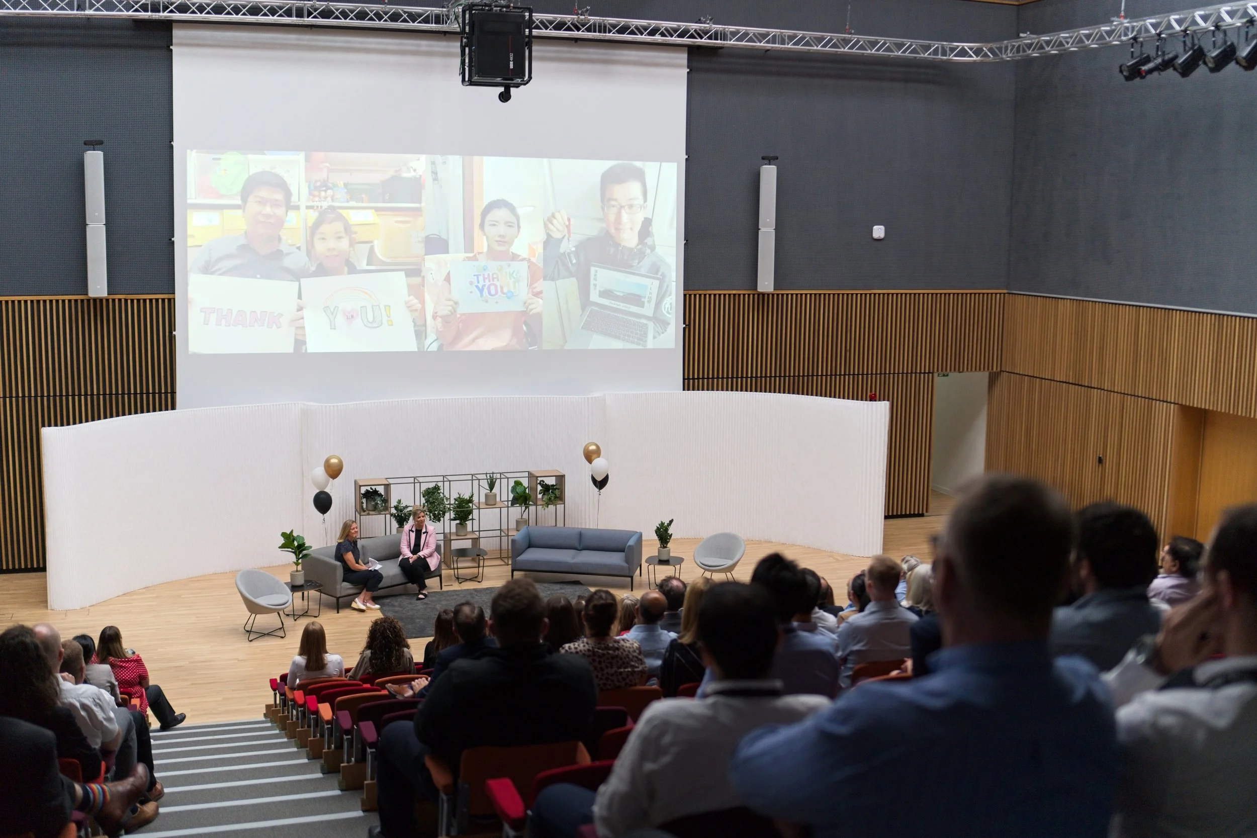 Audience attending a presentation in an auditorium with a stage, two women seated on chairs, listening to a speaker, and a large screen displaying a photo of four people holding signs and a laptop. Event Photography Warwickshire