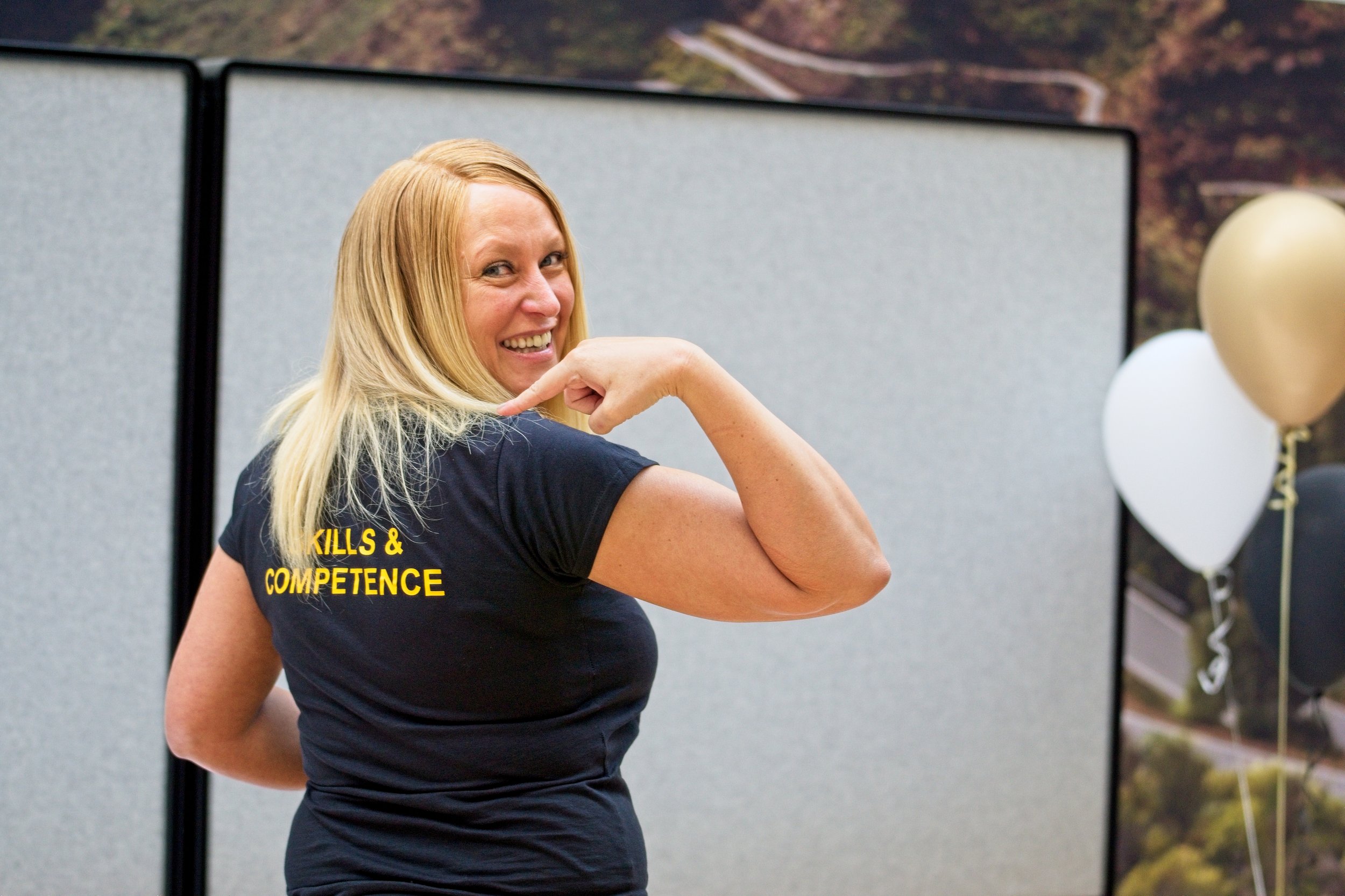 A smiling woman with long blonde hair flexing her arm, wearing a dark T-shirt with yellow text that says 'Skills & Competence', standing indoors with balloons and a scenic backdrop in the background. Event Photography by photographer based in Southam