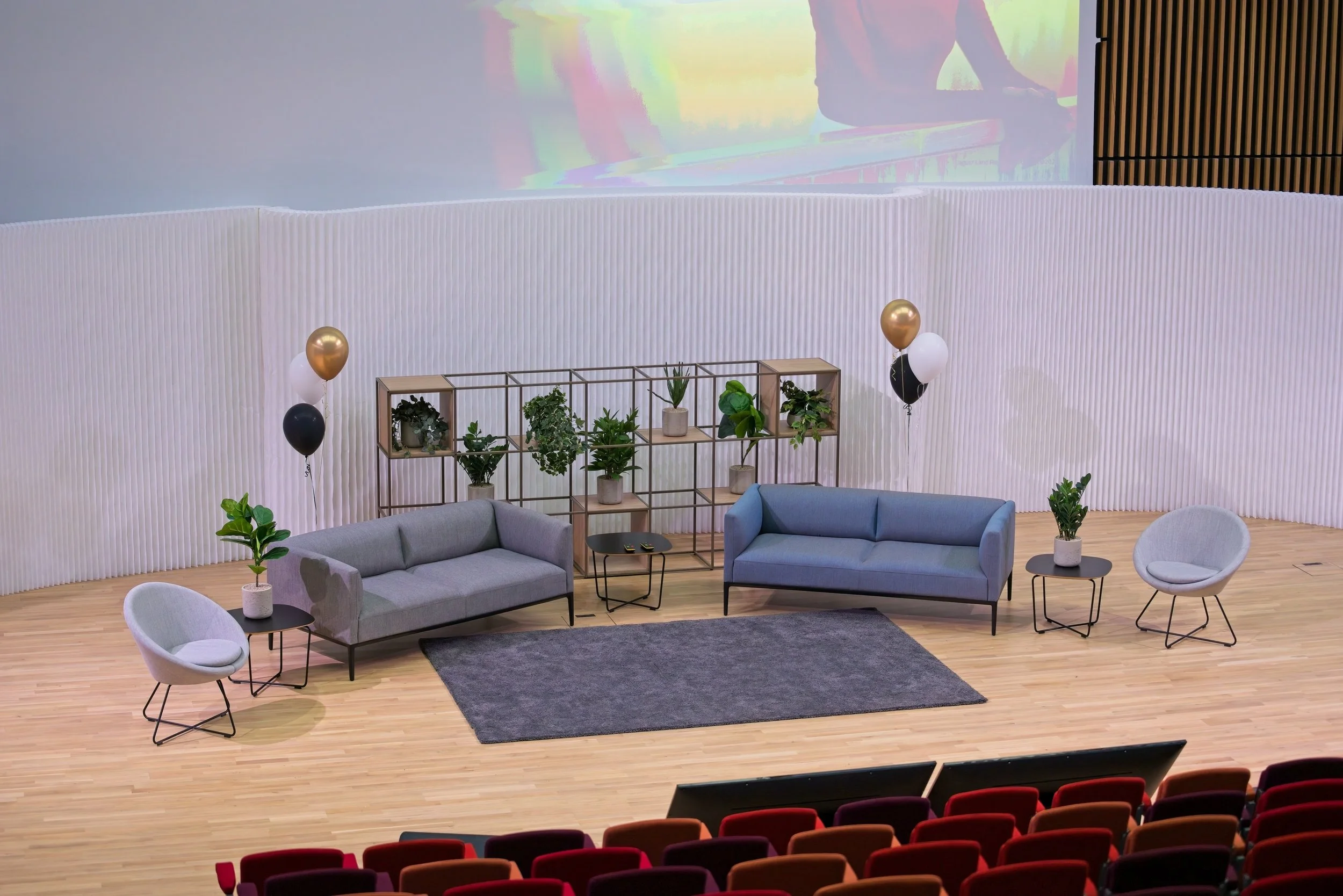 An auditorium stage set with two gray sofas, two round side tables with potted plants, two armchairs, and balloons.  Event Photography Warwickshire