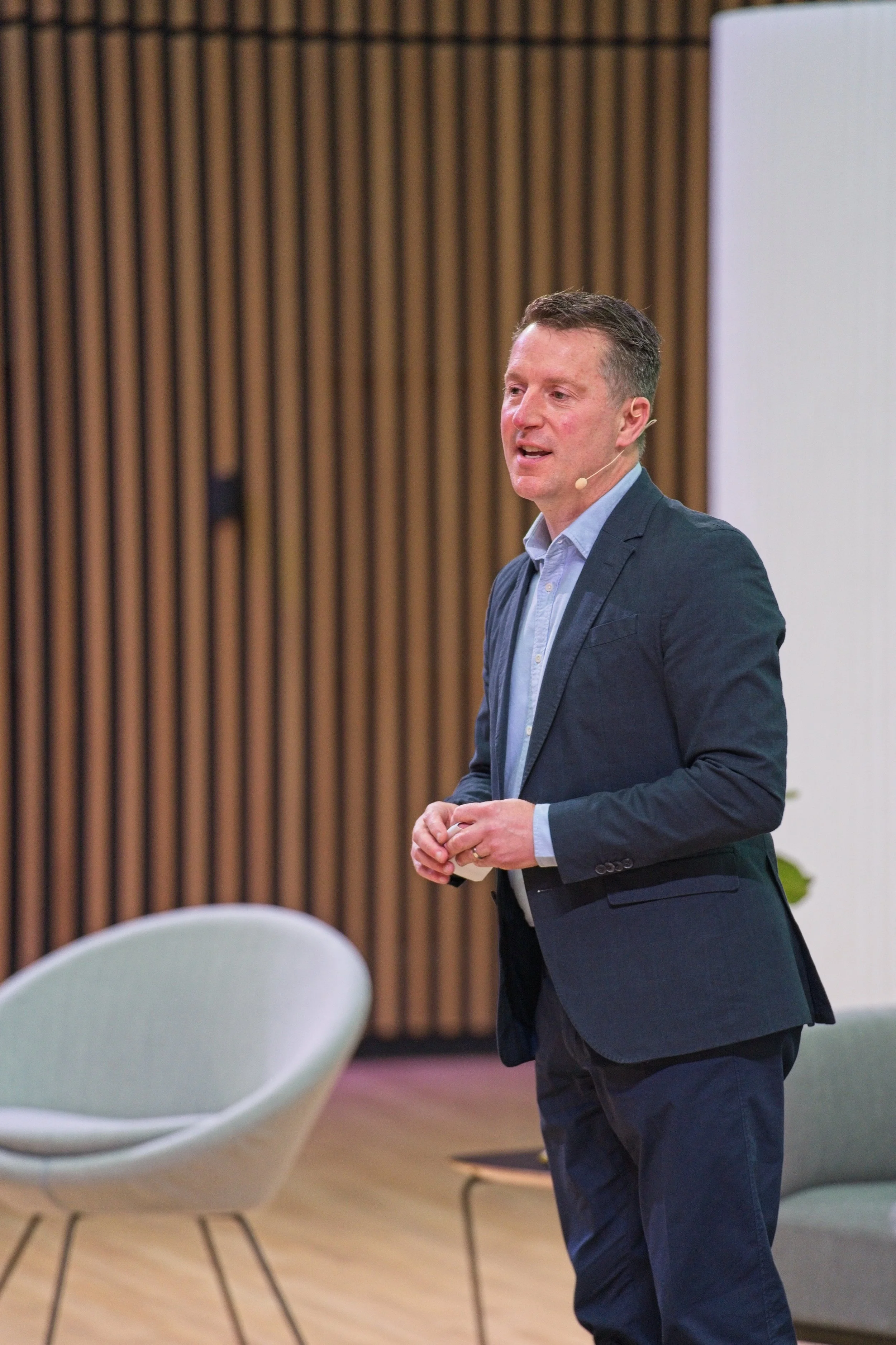 A man in a suit giving a presentation in a modern conference room with wooden panel walls and white chairs. Event Photography by photographer based in Southam, Warwickshire.